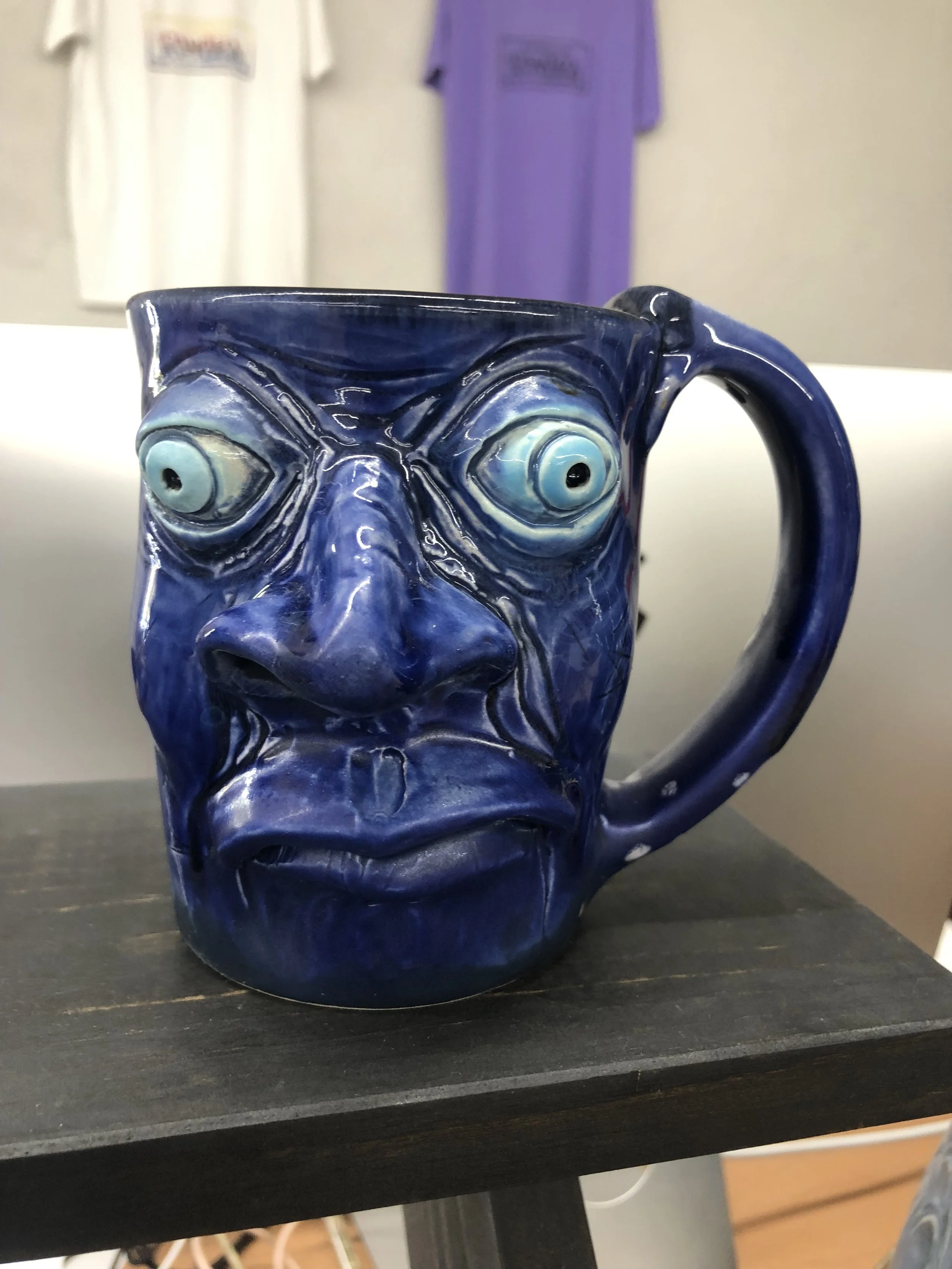 A blue ceramic mug with a face design, featuring large, bulging eyes and exaggerated facial features, placed on a black surface.