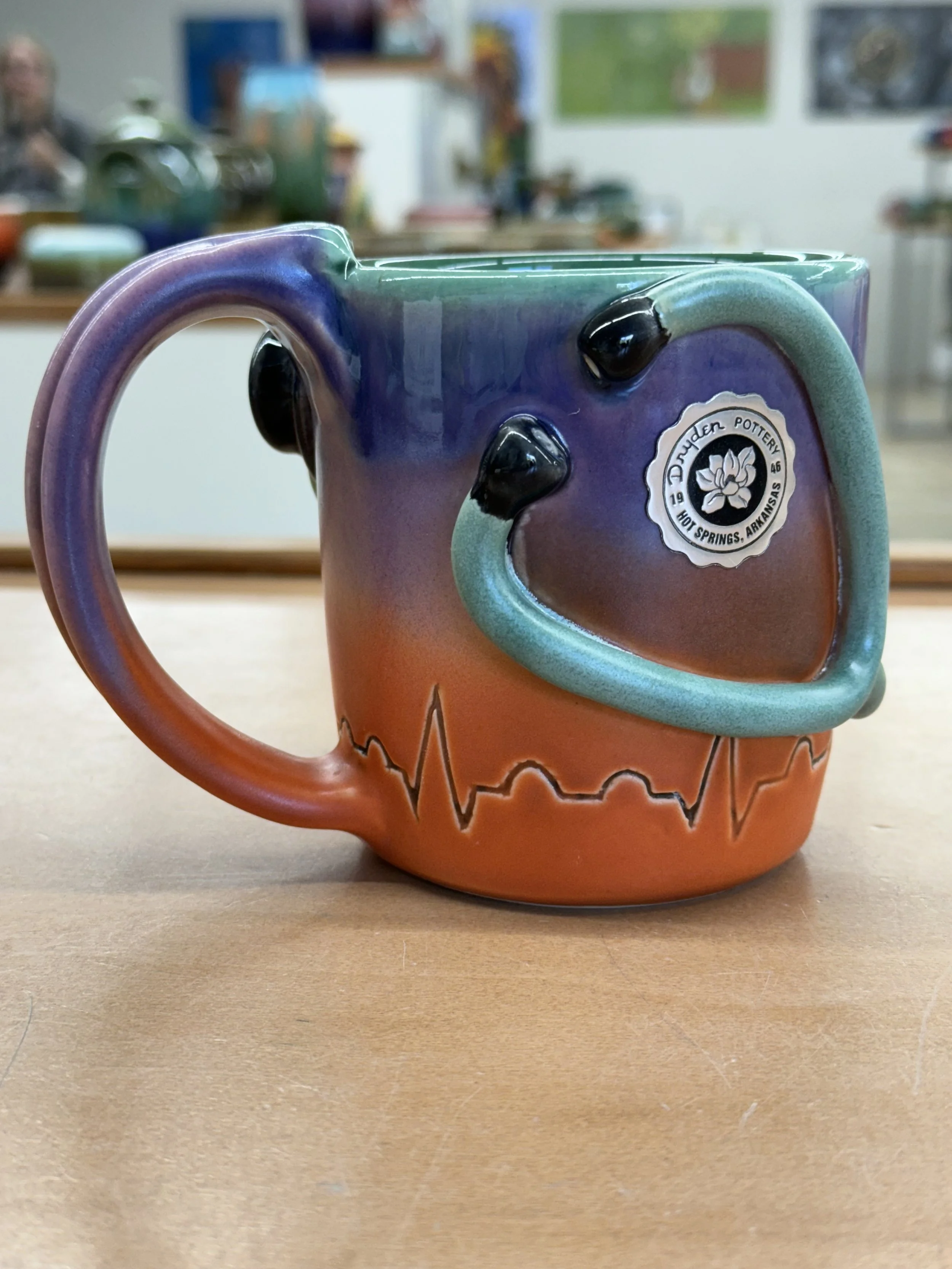 Colorful ceramic coffee mug with a gradient from purple to orange, decorated with black raised knobs and an electrocardiogram pattern around the bottom, displayed on a wooden surface in a pottery studio.