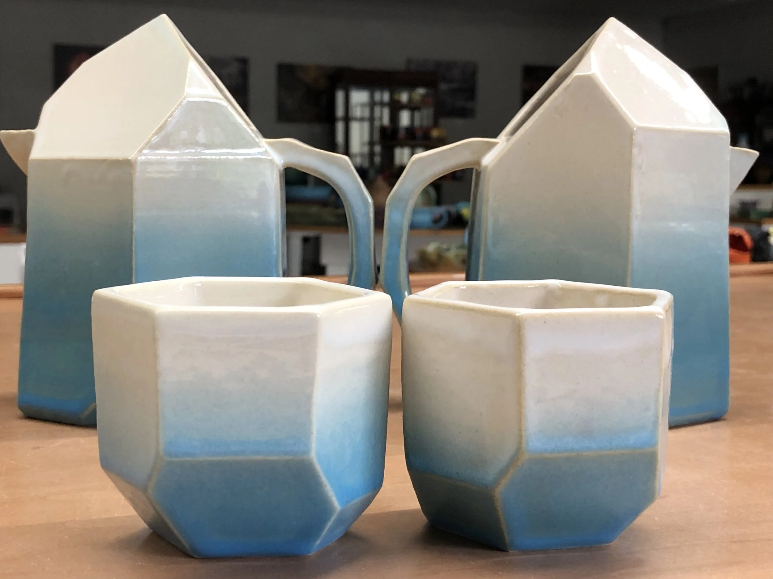 Set of four ceramic mugs and cups with geometric faceted design, glazed in a gradient of white to blue, arranged on a wooden surface.