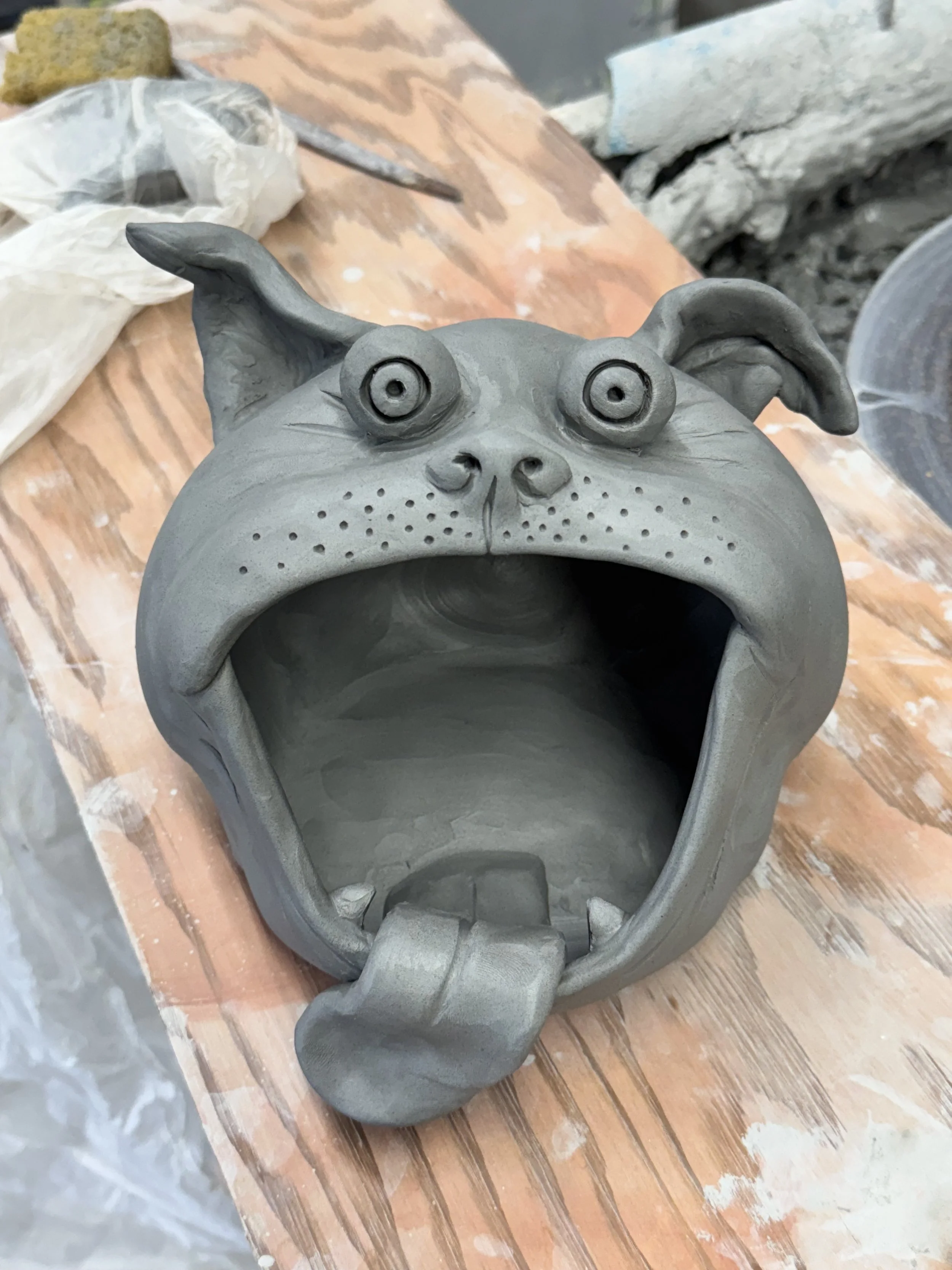 Clay sculpture of a humorous dog head with large open mouth, tongue sticking out, big eyes, and floppy ears, on a wooden work surface.