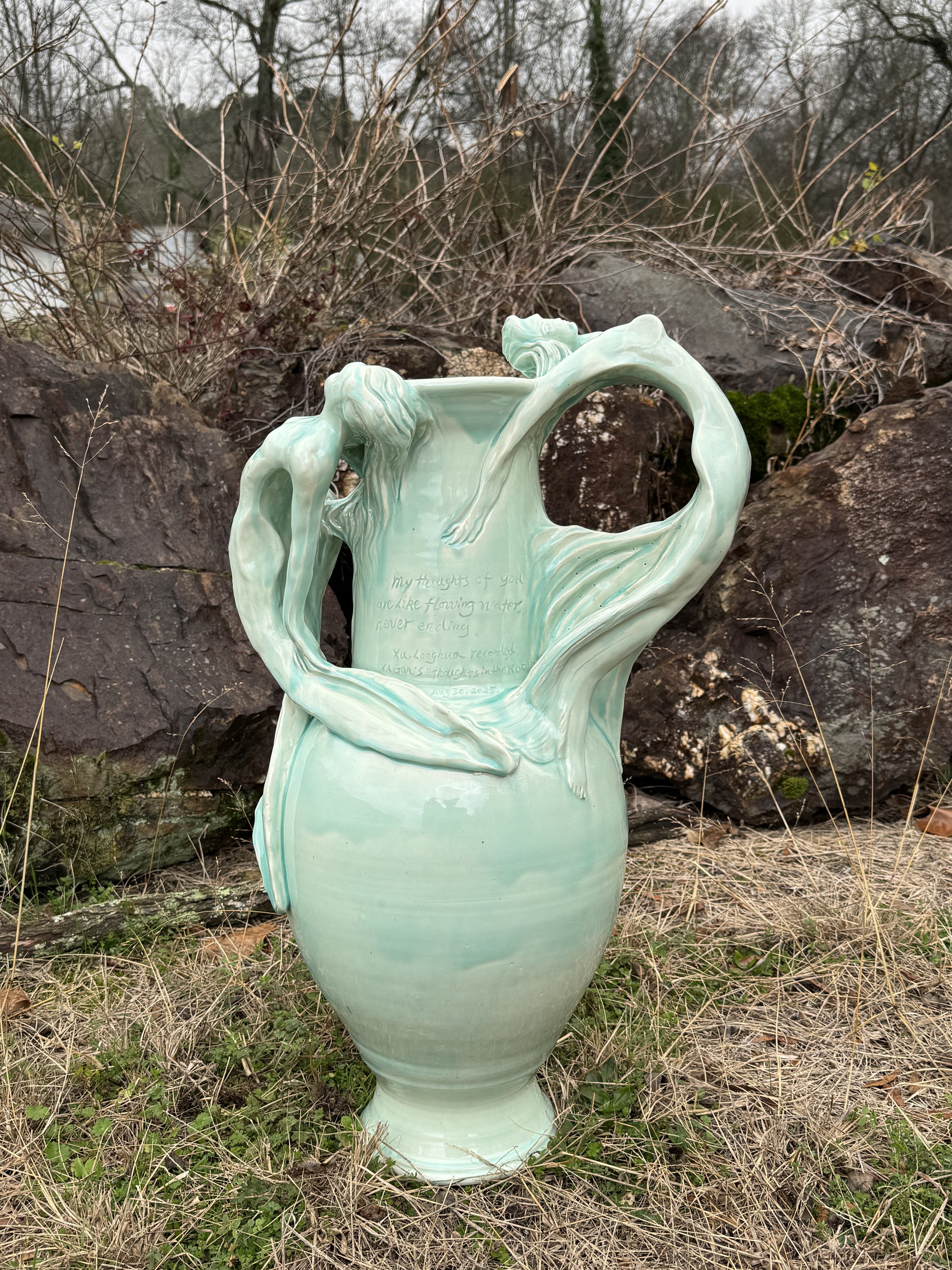 A green ceramic sculpture of three intertwined female figures forming a heart shape, placed outdoors among rocks and dry grass.