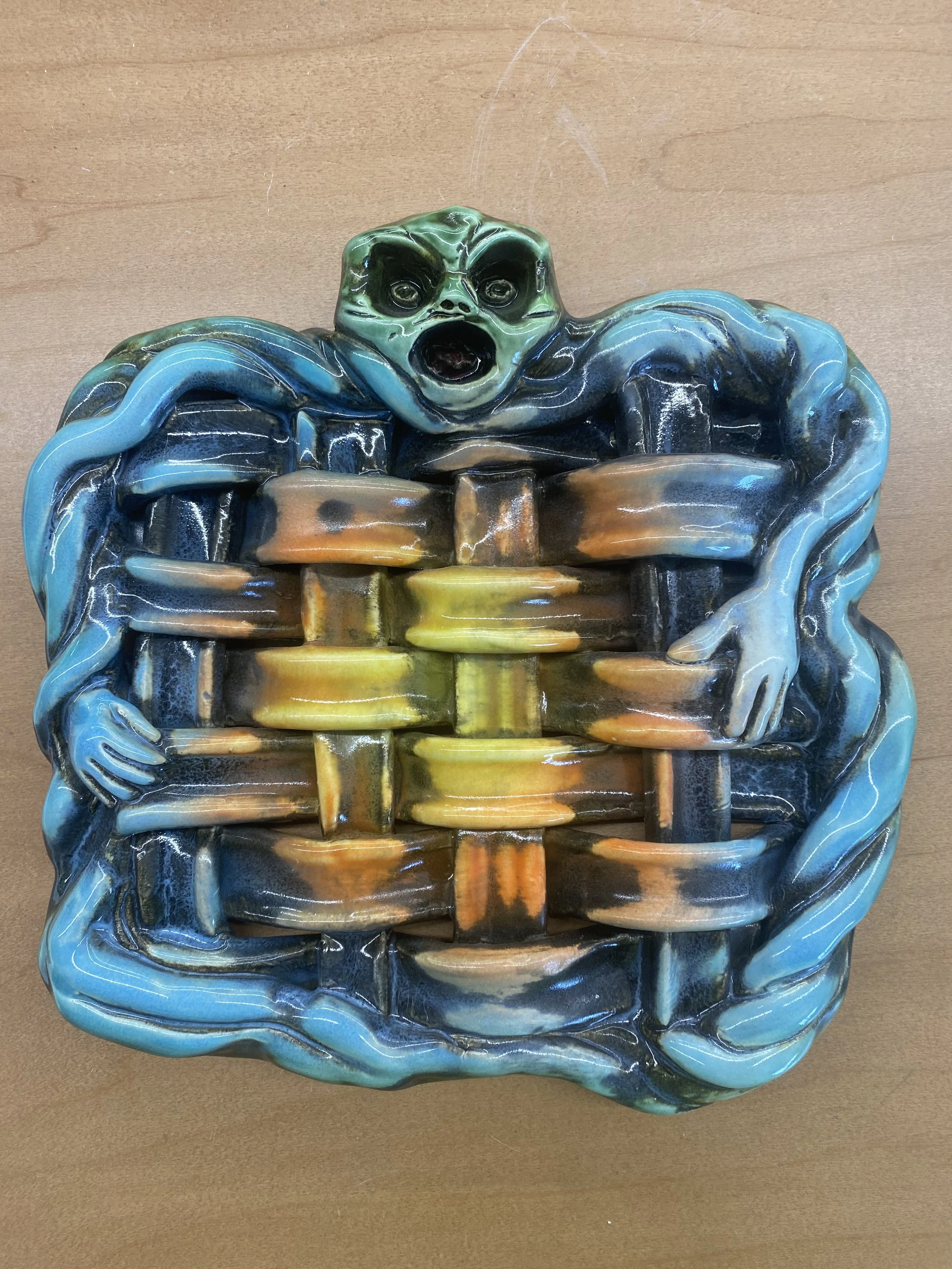 Colorful ceramic sculpture depicting a creature with a human-like face and arms, emerging from a woven basket.