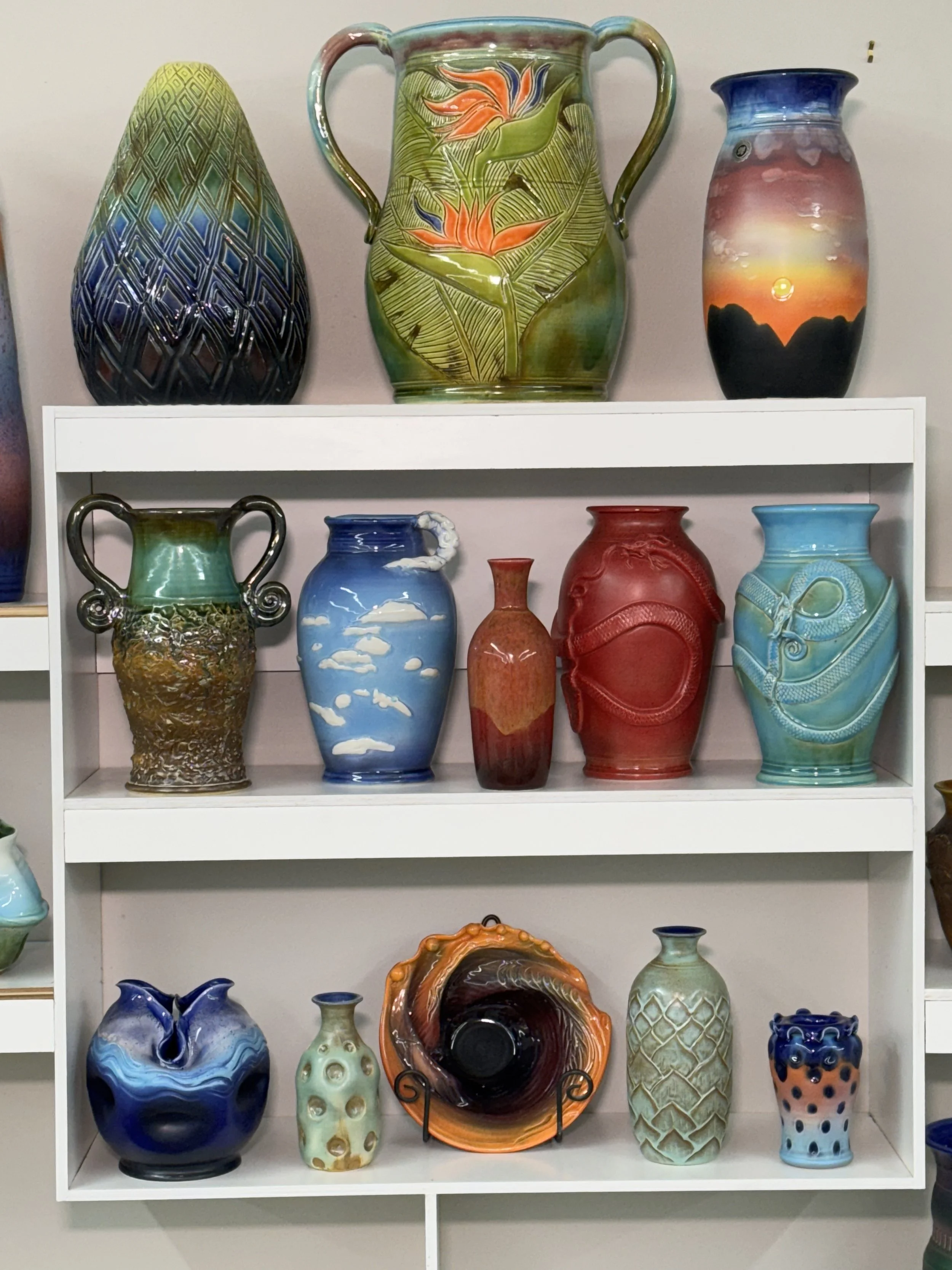 Display of various colorful decorative vases on white shelves, featuring different shapes, sizes, and artistic designs.