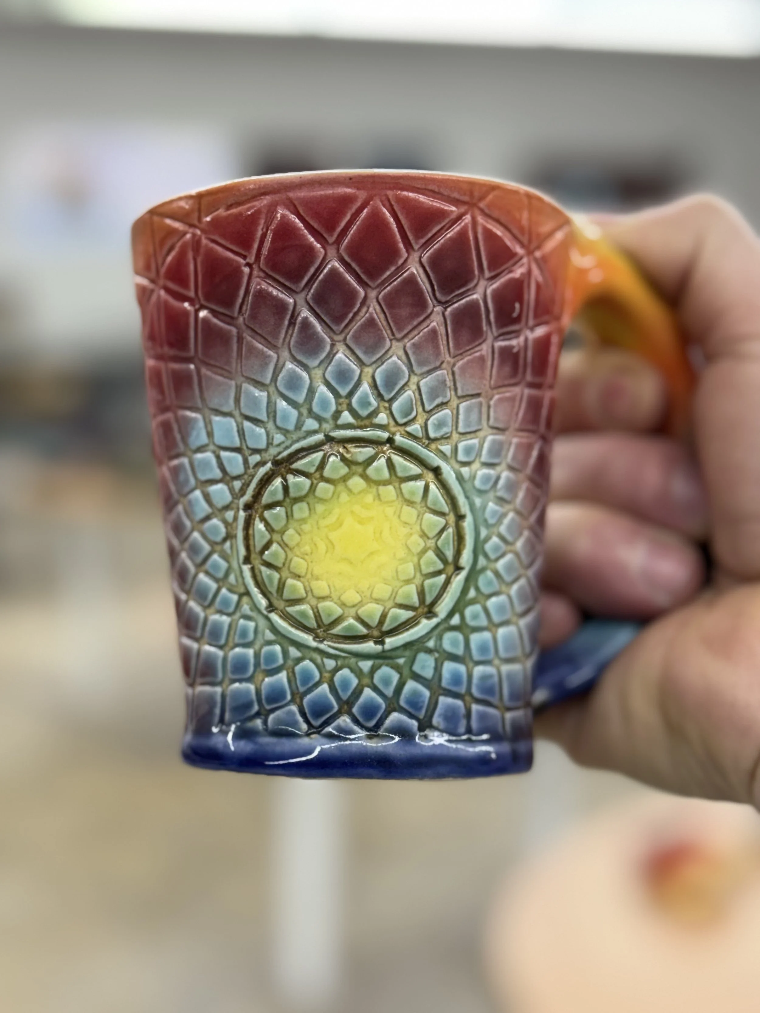 Colorful glass mug with geometric pattern, held by a person's hand, blurry background.