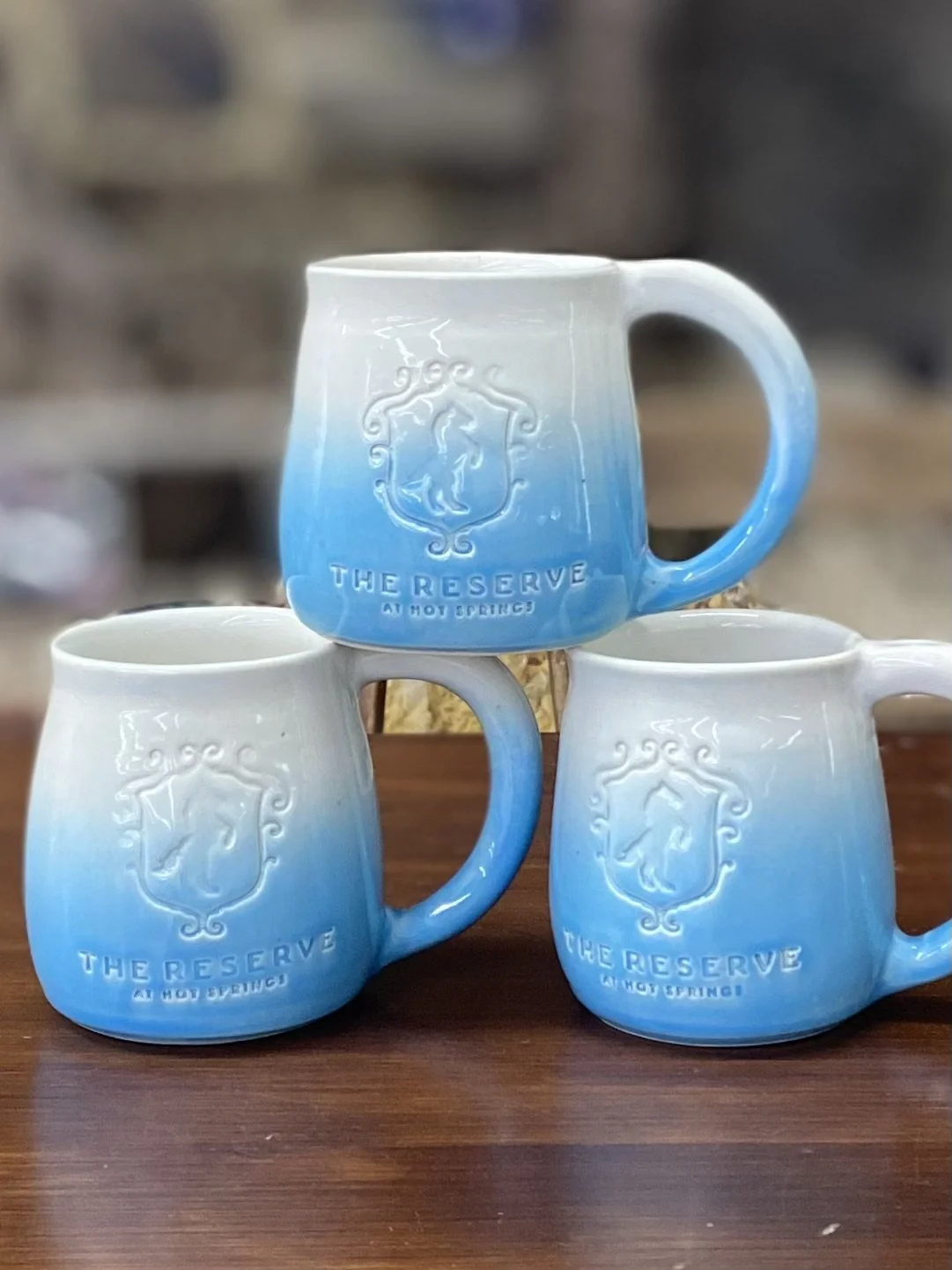 Three ceramic mugs with a blue and white gradient design, embossed with a crest and the words "The Reserve at Hot Springs," arranged on a wooden surface.