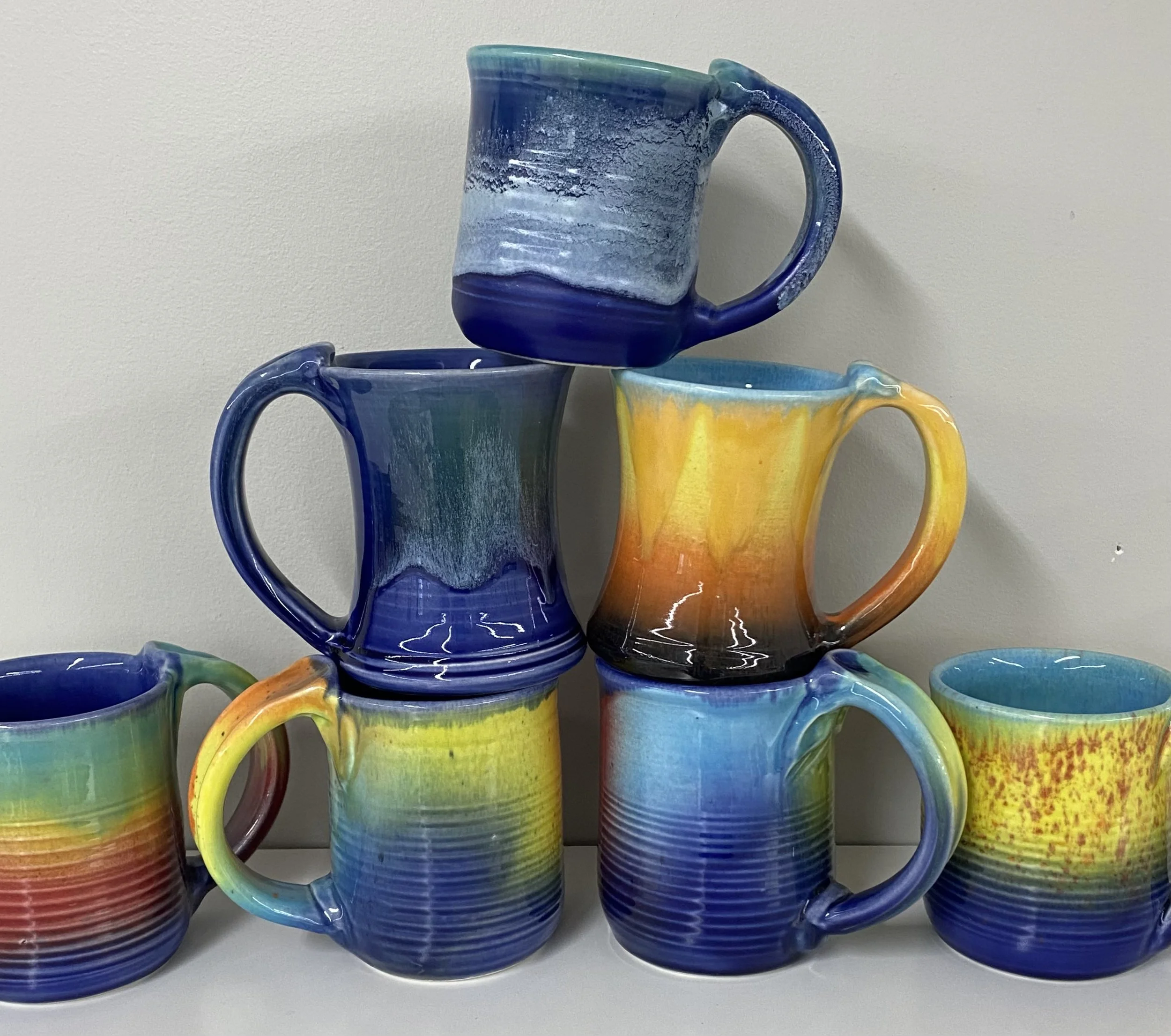 Six colorful ceramic mugs, three stacked in a pyramid shape on a light-colored surface against a plain background, featuring vibrant rainbow-like and ocean-blue glaze patterns.
