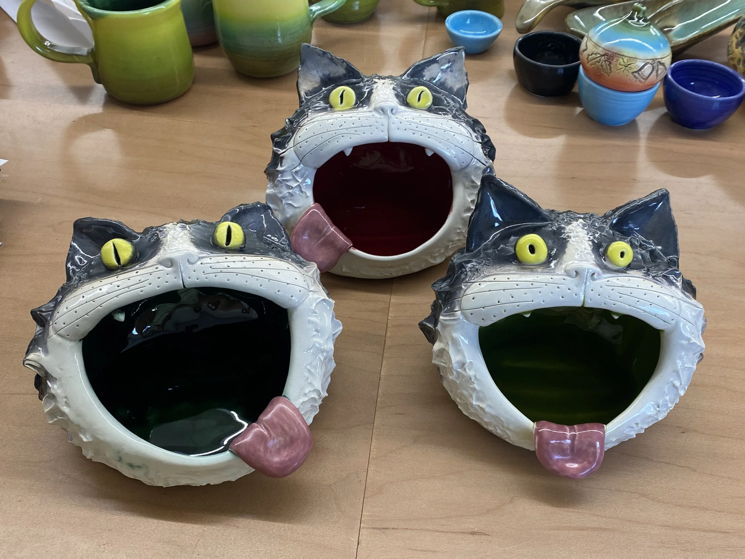 Three ceramic cat face-shaped vessels with wide open mouths, yellow eyes, pink tongues, and white fur, placed on a wooden surface with various colorful cups and mugs in the background.