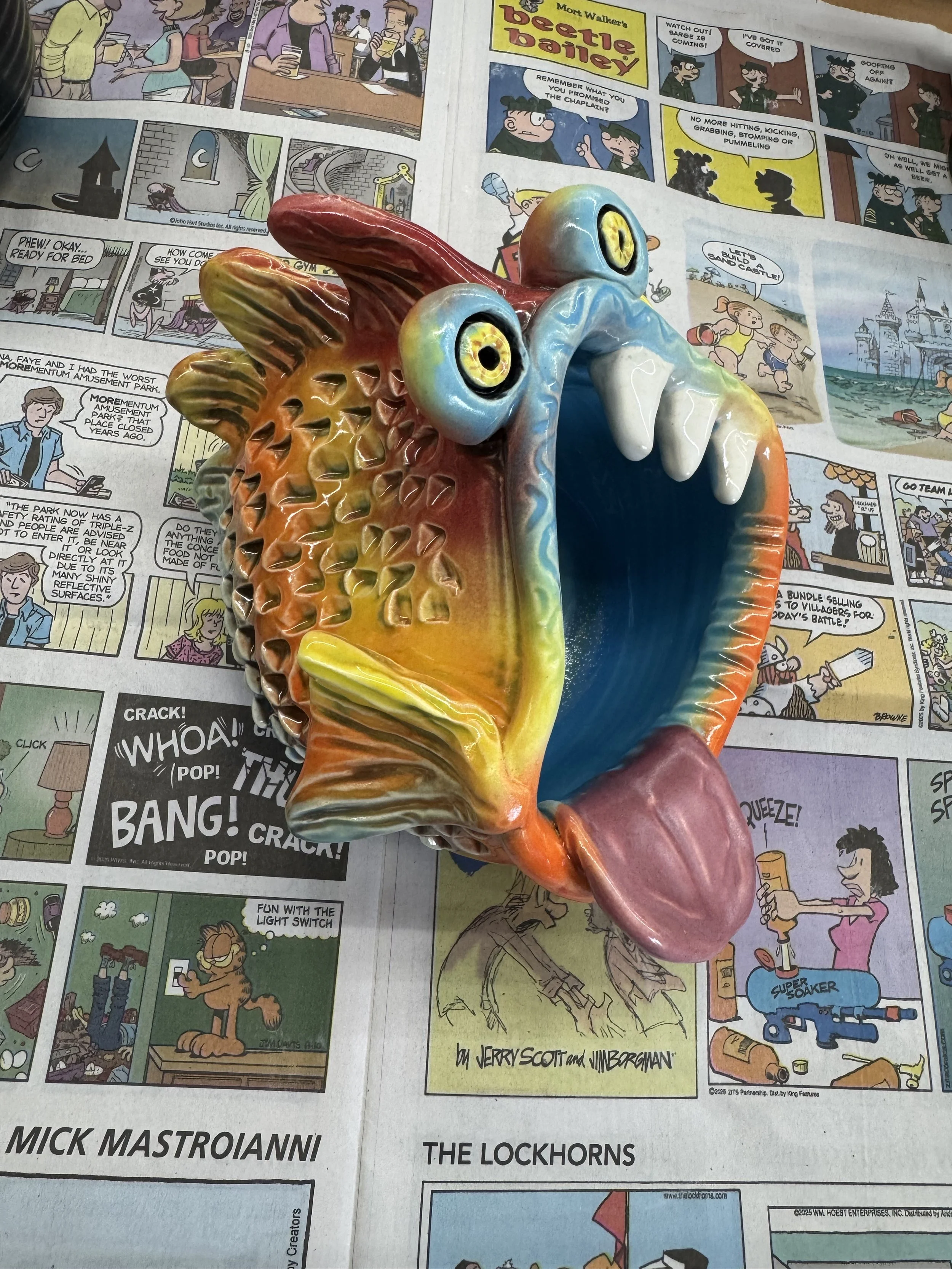 Colorful ceramic fish sculpture with wide open mouth, big eyes, and detailed scales, placed on a newspaper page with comic strips.