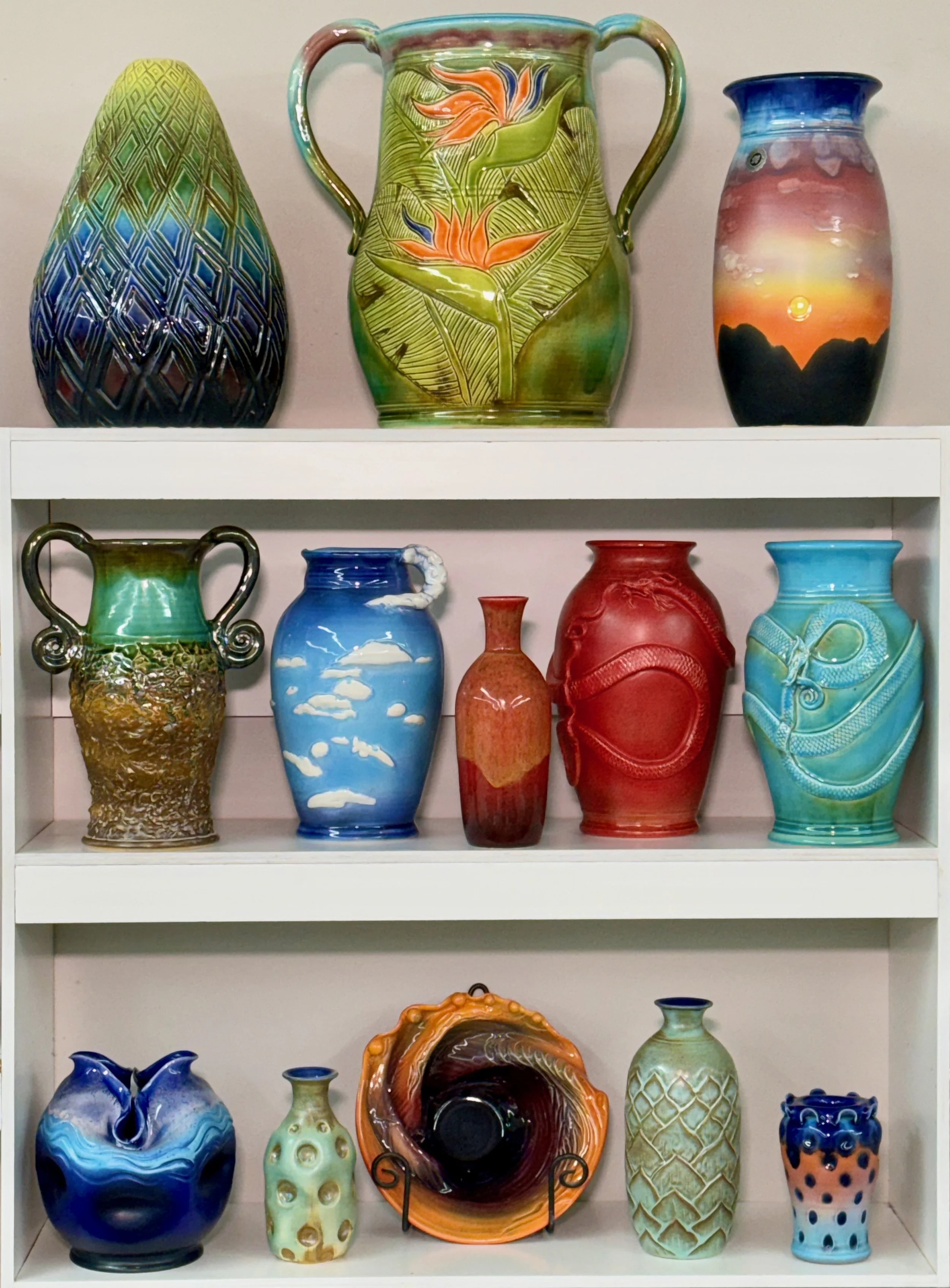 A collection of decorative glass vases and pottery arranged on white shelves, featuring various colors, shapes, and artistic designs.
