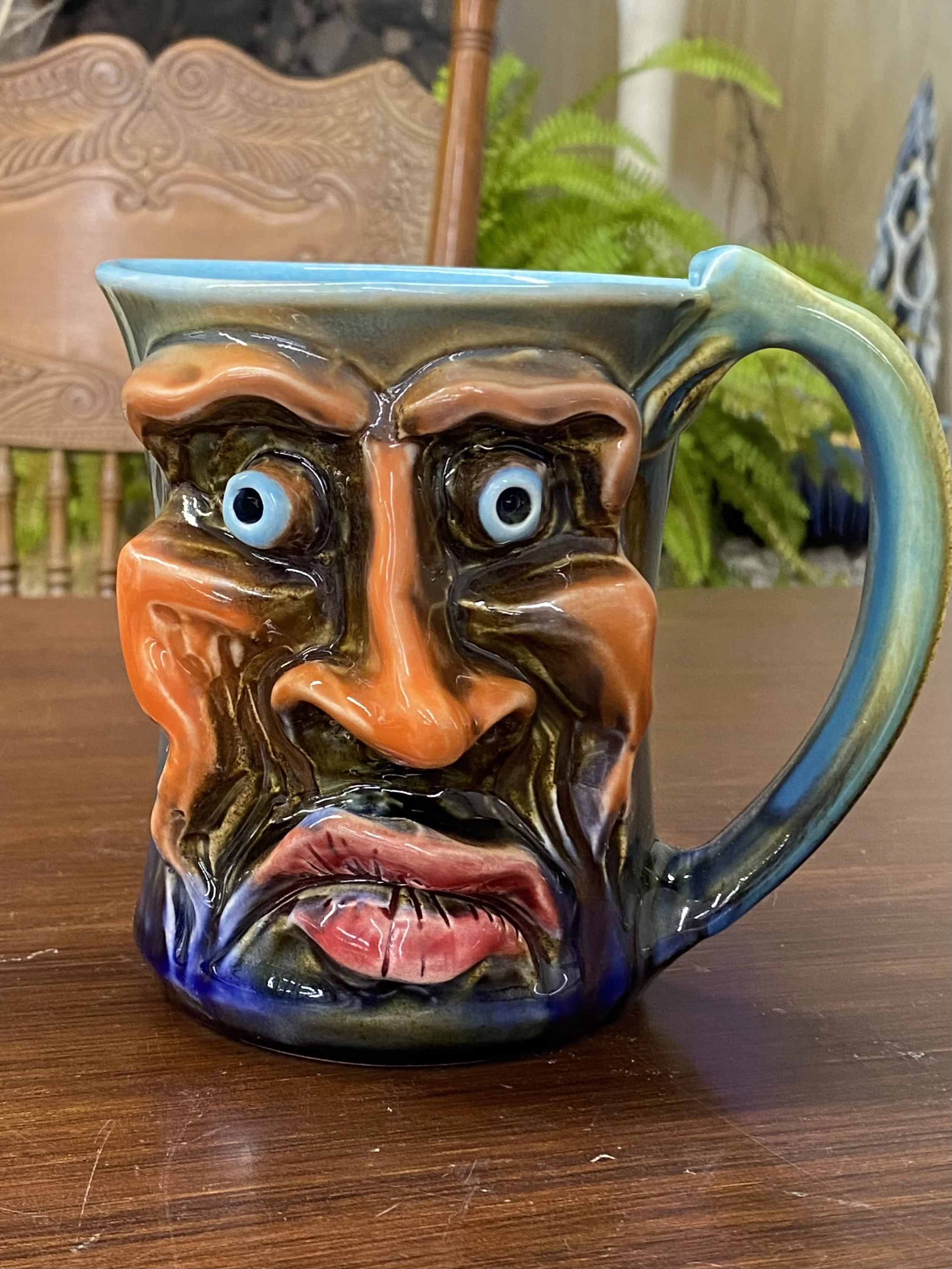 A ceramic mug with a distorted human face design, featuring exaggerated features including eyes, nose, mouth, and brows, in vibrant colors.