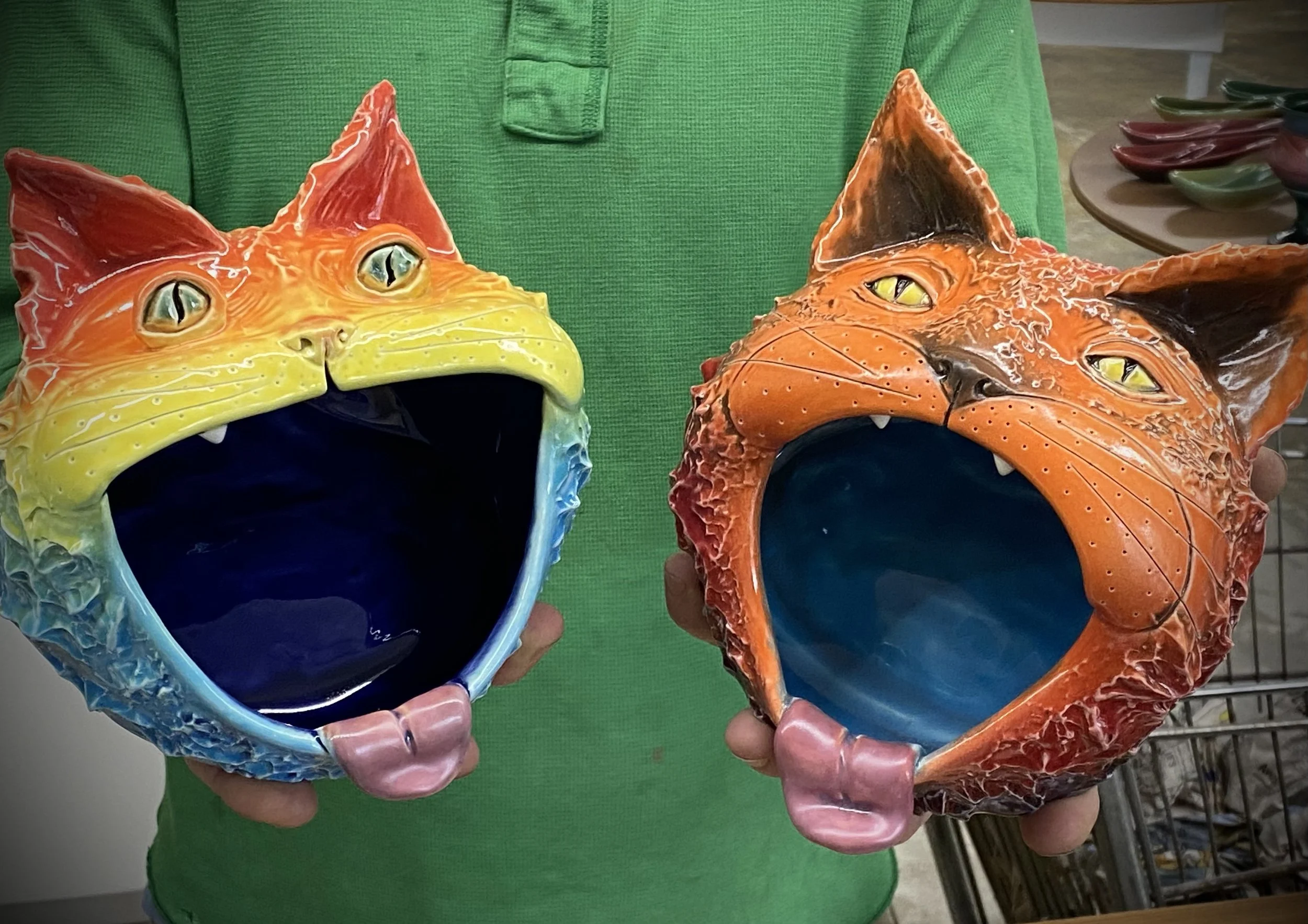 Person in green shirt holding colorful ceramic cat face bowls, one with rainbow colors and the other with orange and black, both with wide open mouths.