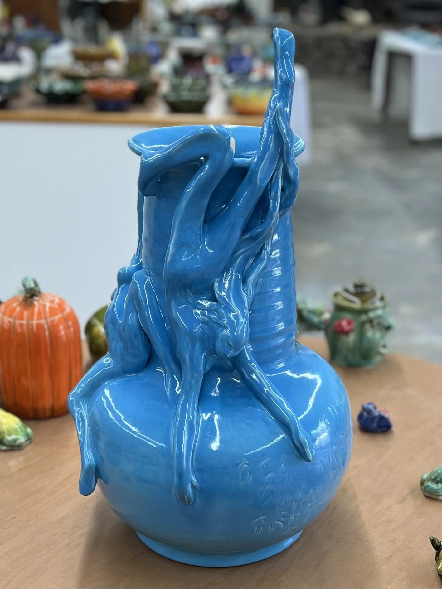 Blue ceramic vase with ornate female figures relief design, displayed on a wooden surface with other colorful pottery in the background.