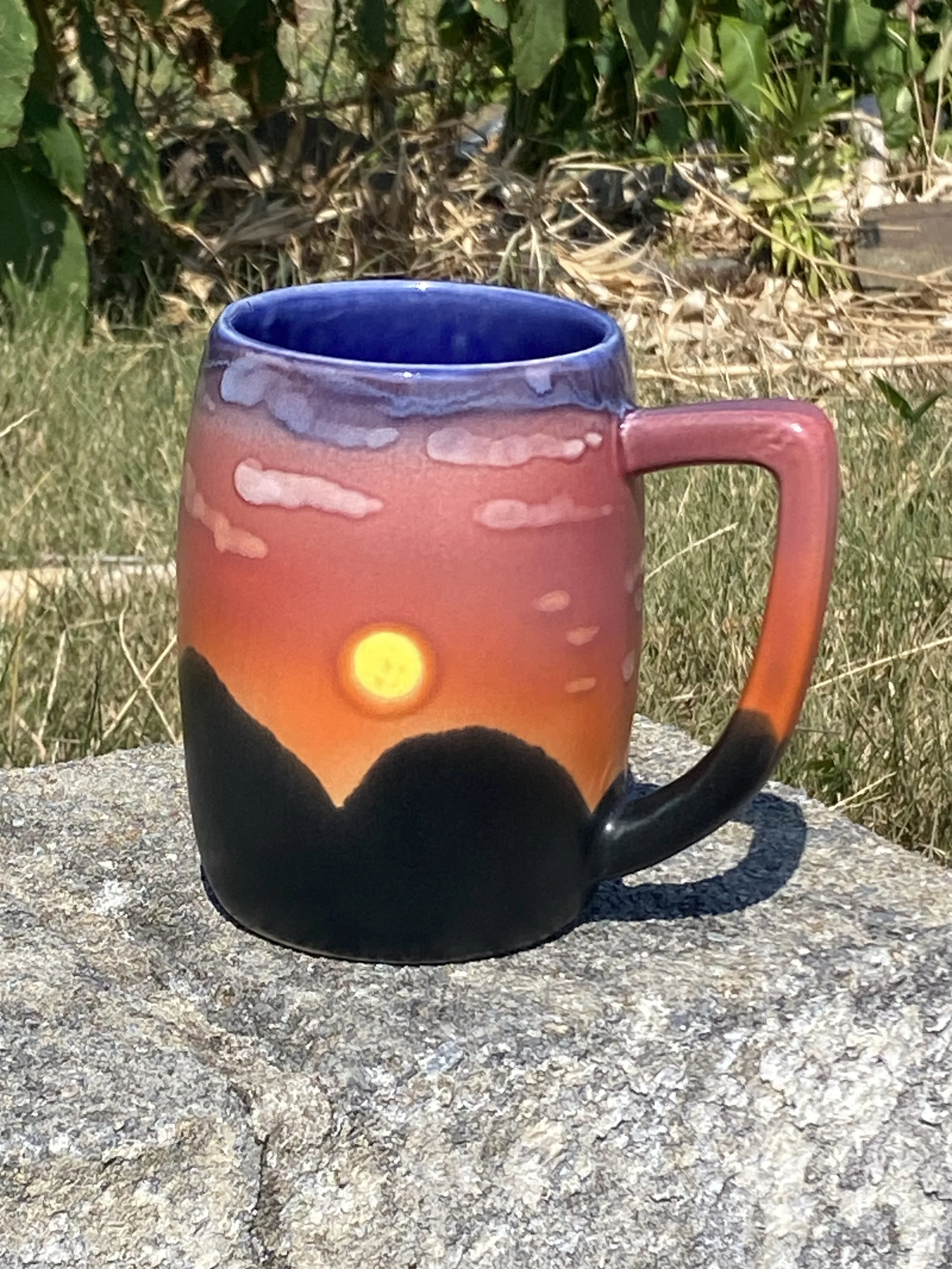A ceramic mug with a sunset scene painted on it, featuring a yellow sun setting behind dark mountains, with a pink and purple sky and gray clouds, placed on a stone surface outdoors.
