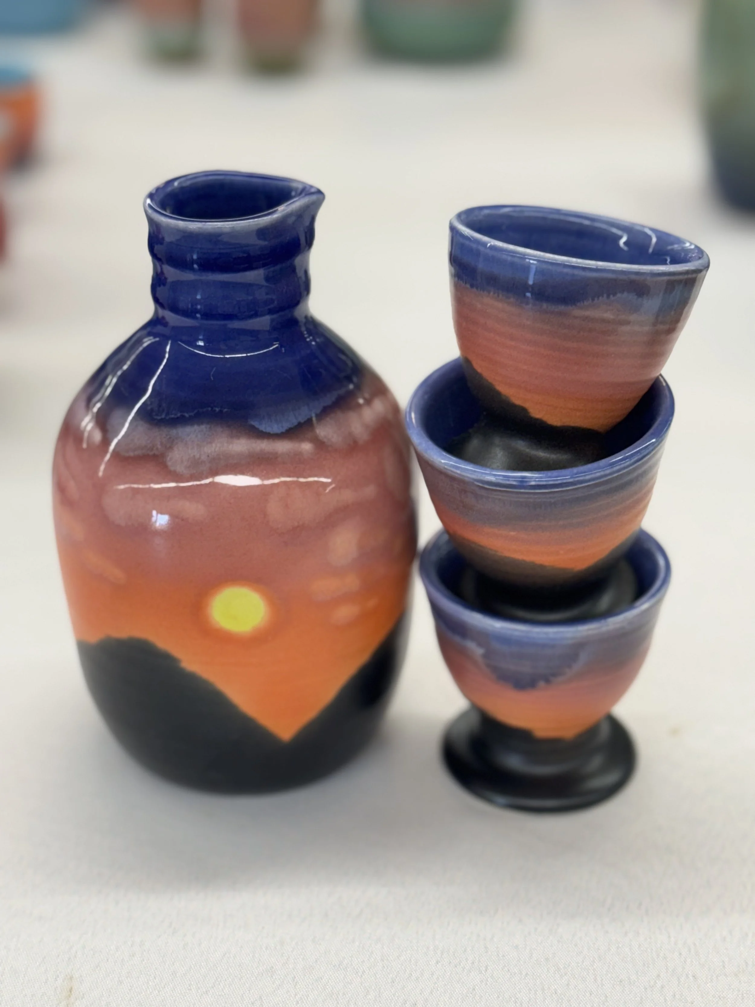 Ceramic vase and three small cups with sunset and mountain landscape painting, placed on a white surface.