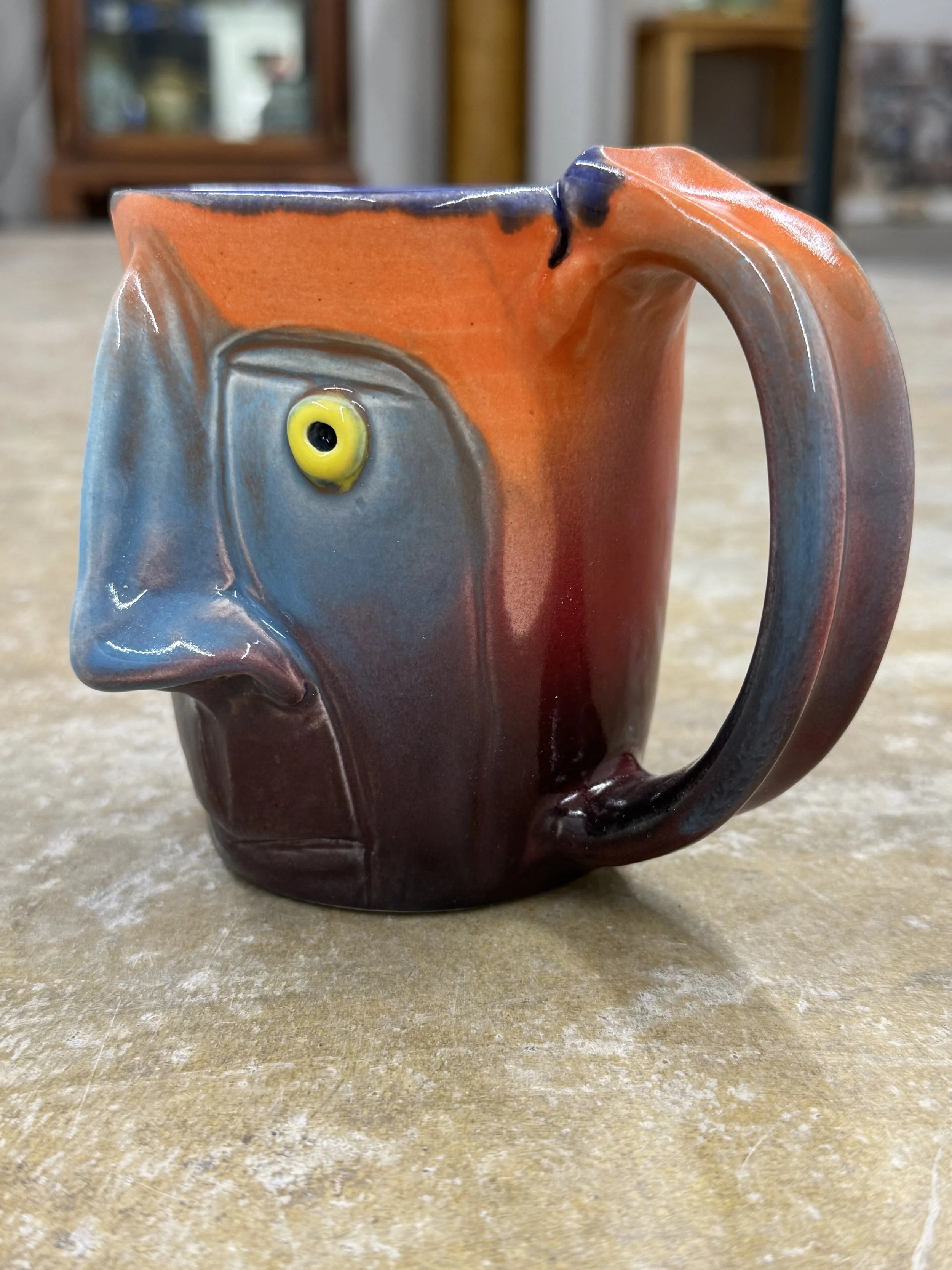 A ceramic mug with a face and a nose, painted in orange, blue, and black, with a yellow and black eye, sitting on a wooden surface.