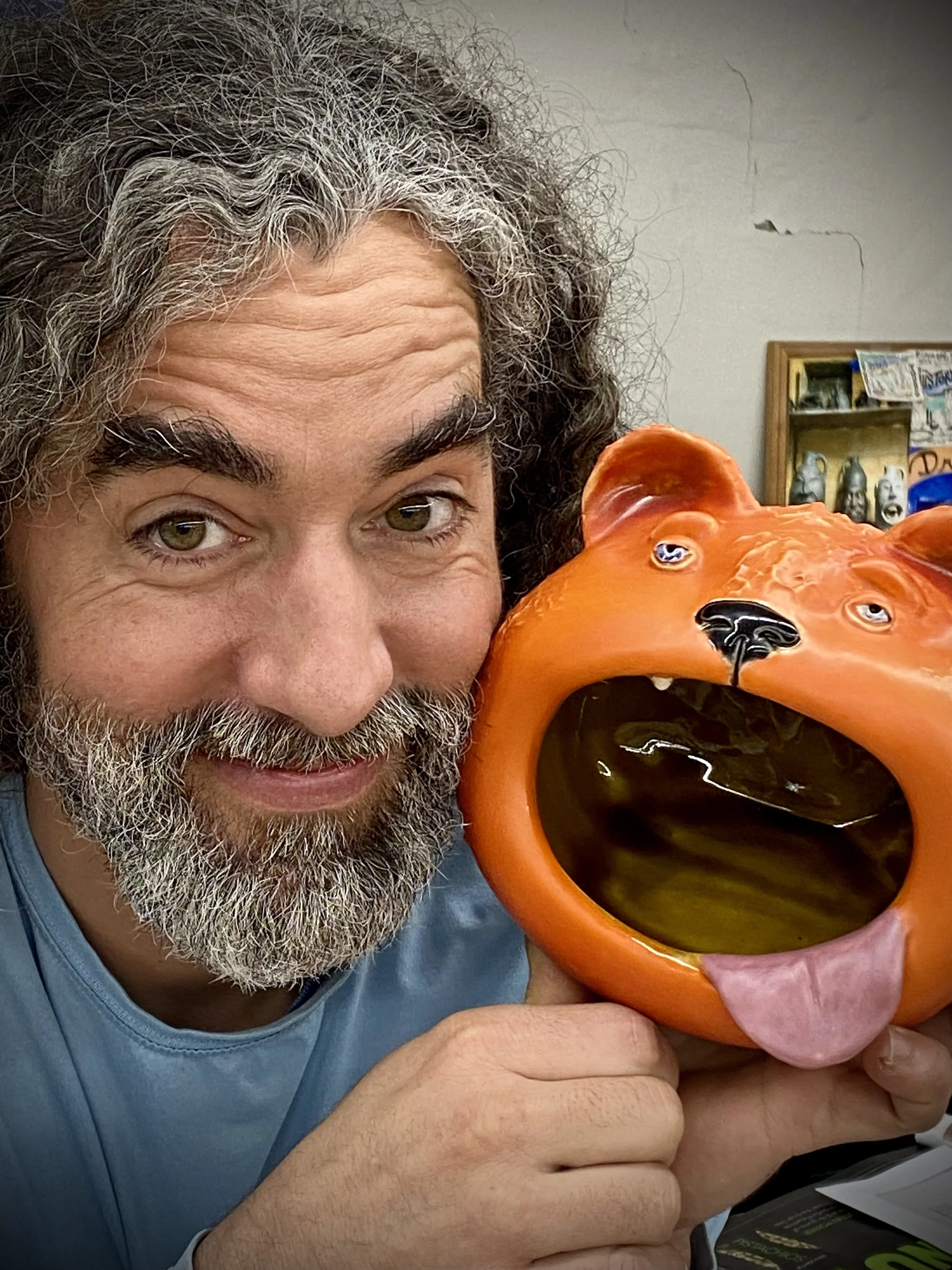 A man with gray curly hair and a beard holding an orange ceramic bear head-shaped sculpture with its mouth open, tongue out, and eyes looking forward.