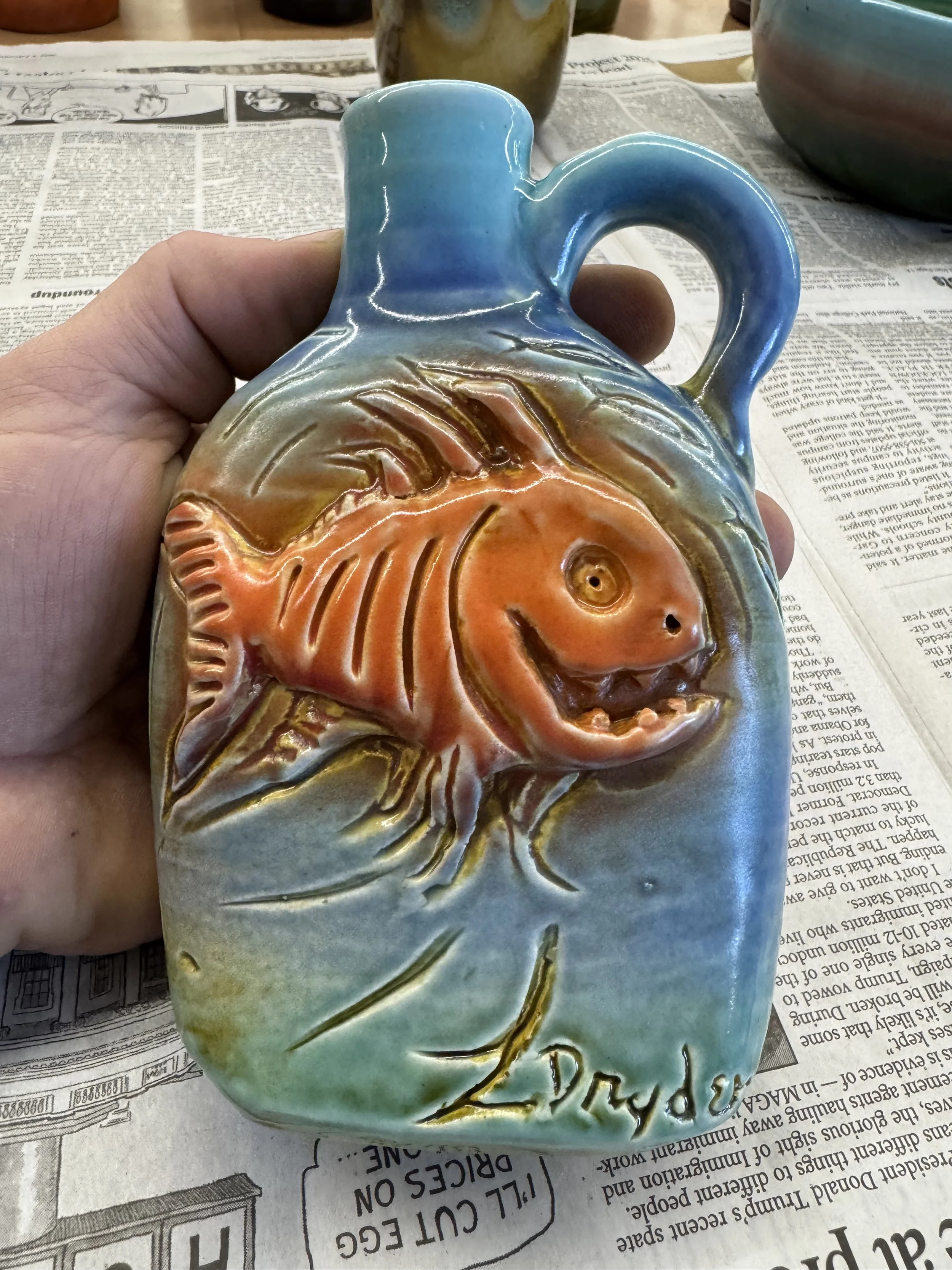 Colorful ceramic vase with a fish design, featuring orange, blue, and green hues, placed on newspaper.