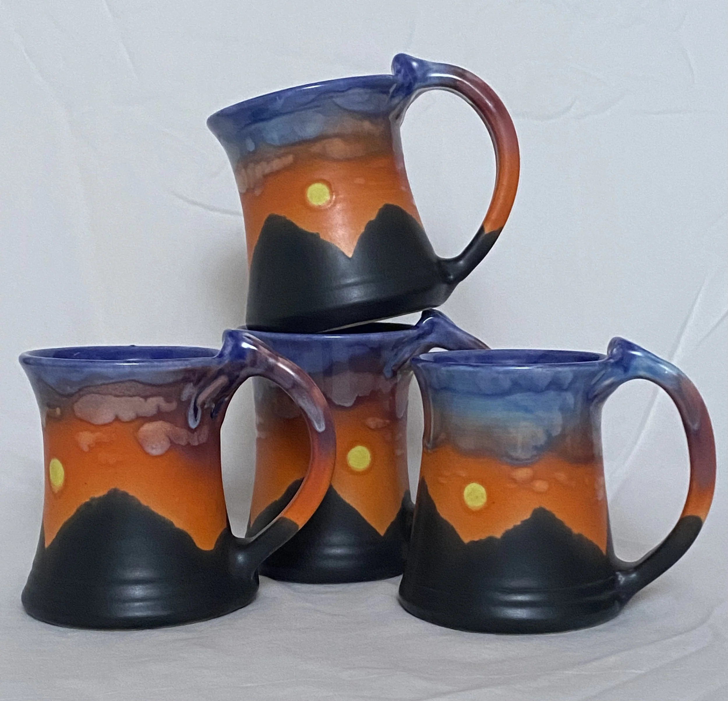 Four ceramic mugs painted with mountain and sunset scenery, featuring orange, black, blue, and yellow colors.