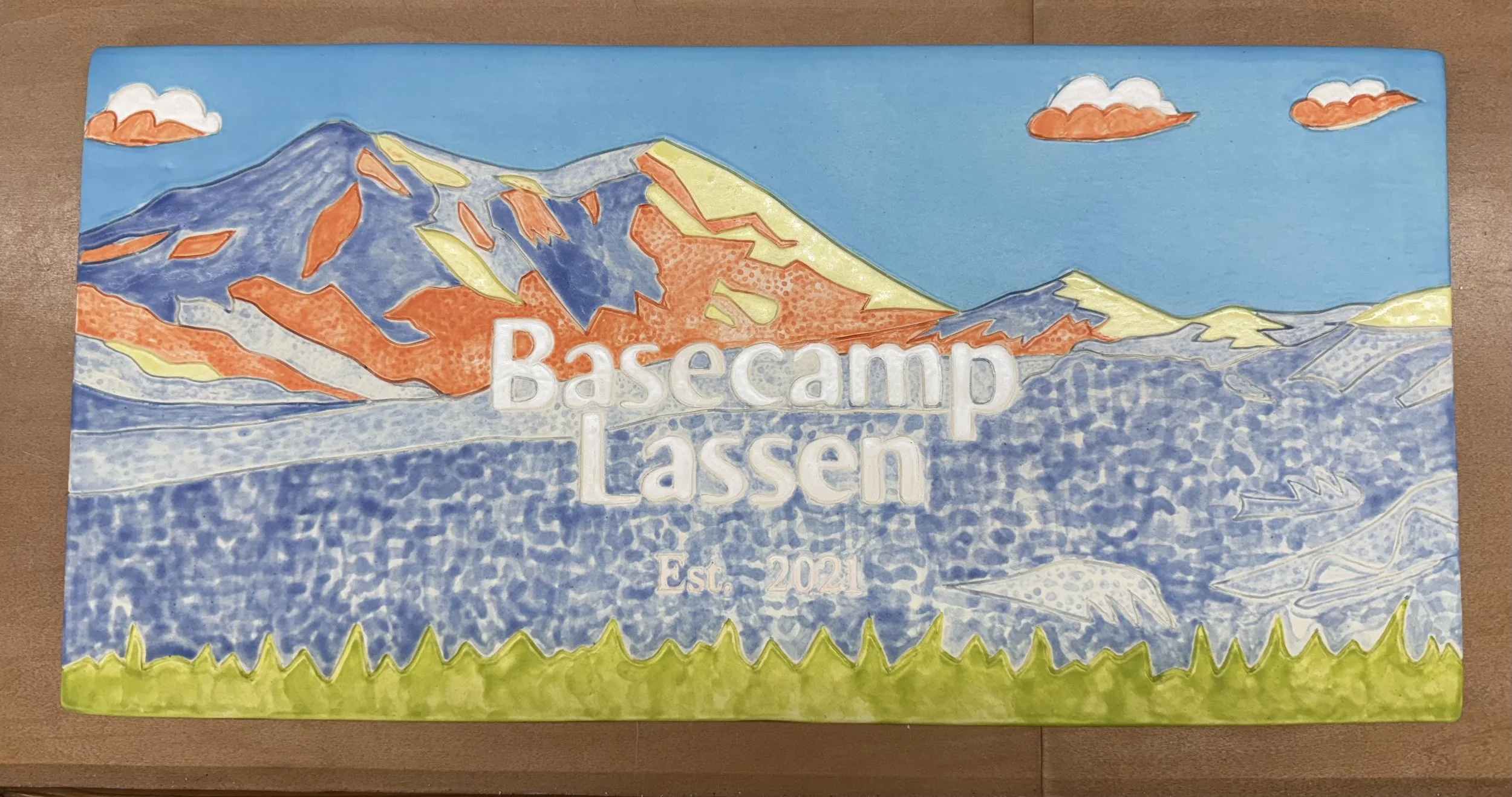 Colorful drawing of mountains, sky, clouds, and water with the text 'Basecamp Lassen Est. 2021' in the center.