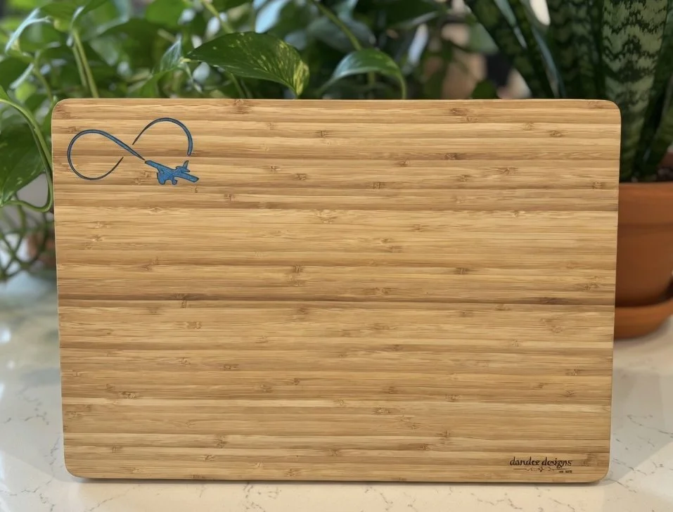 A rectangular bamboo cutting board with a small blue and black fish-shaped charm attached to a string in the top left corner, with green houseplants in the background.