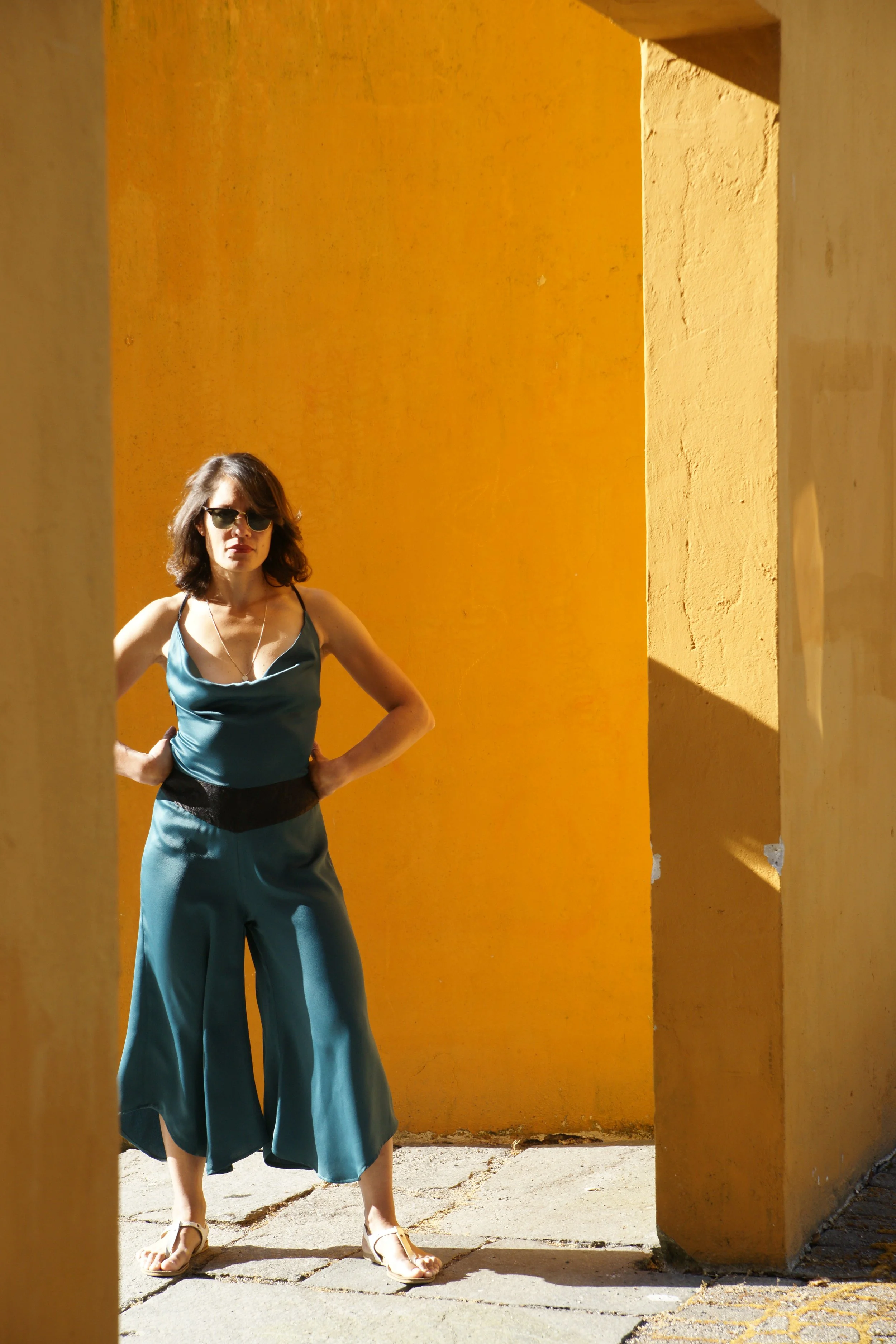 A woman with shoulder-length brown hair wearing sunglasses, a teal satin top, matching satin wide-leg pants, and white sandals stands with her hands on her hips, posing against a vibrant orange wall framed by two yellowish columns.