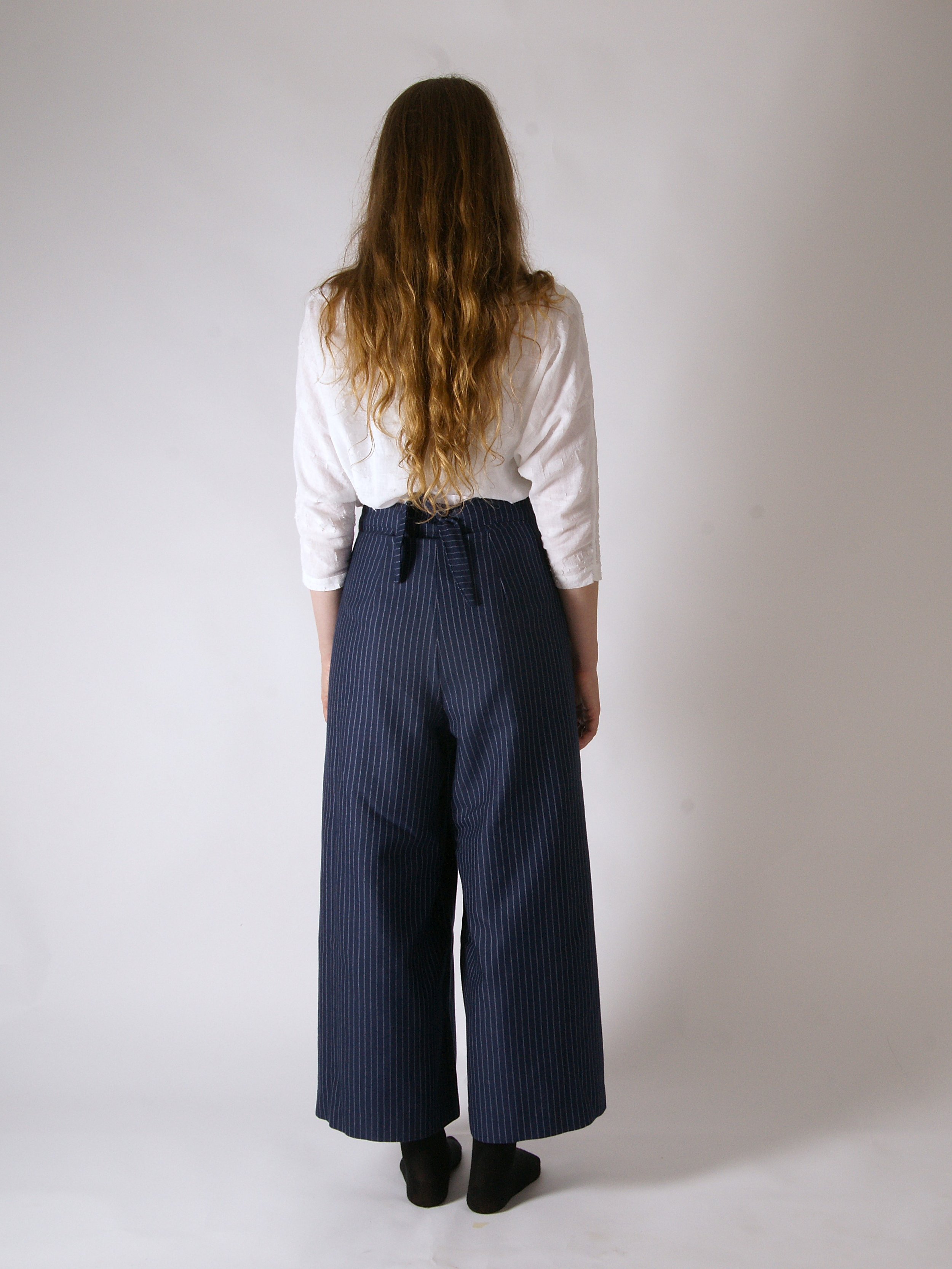 A woman with long, wavy brown hair is standing facing away from the camera against a plain white background. She is wearing a white blouse and navy blue, high-waisted, wide-leg pants with white pinstripes and a tied waist. She is also wearing black s