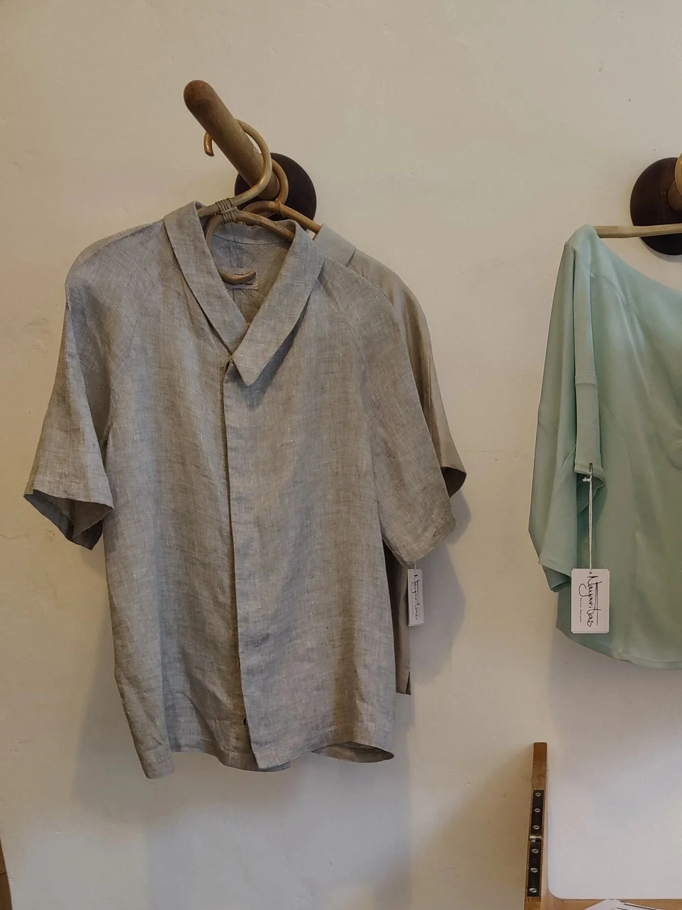 A beige short-sleeved shirt hanging on a wooden hanger against a plain wall, with a tag attached, and a pale green garment to the right also hanging on a hanger.