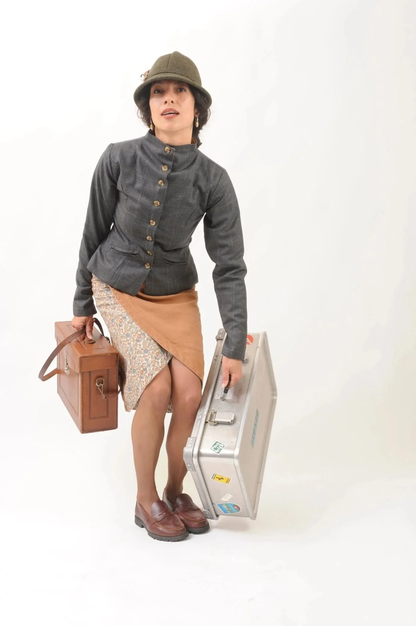 A woman dressed in vintage style clothing, including a gray tailored jacket with gold buttons, a brown skirt, and brown shoes, holding a brown leather suitcase and standing next to a silver metal briefcase, against a plain white background.