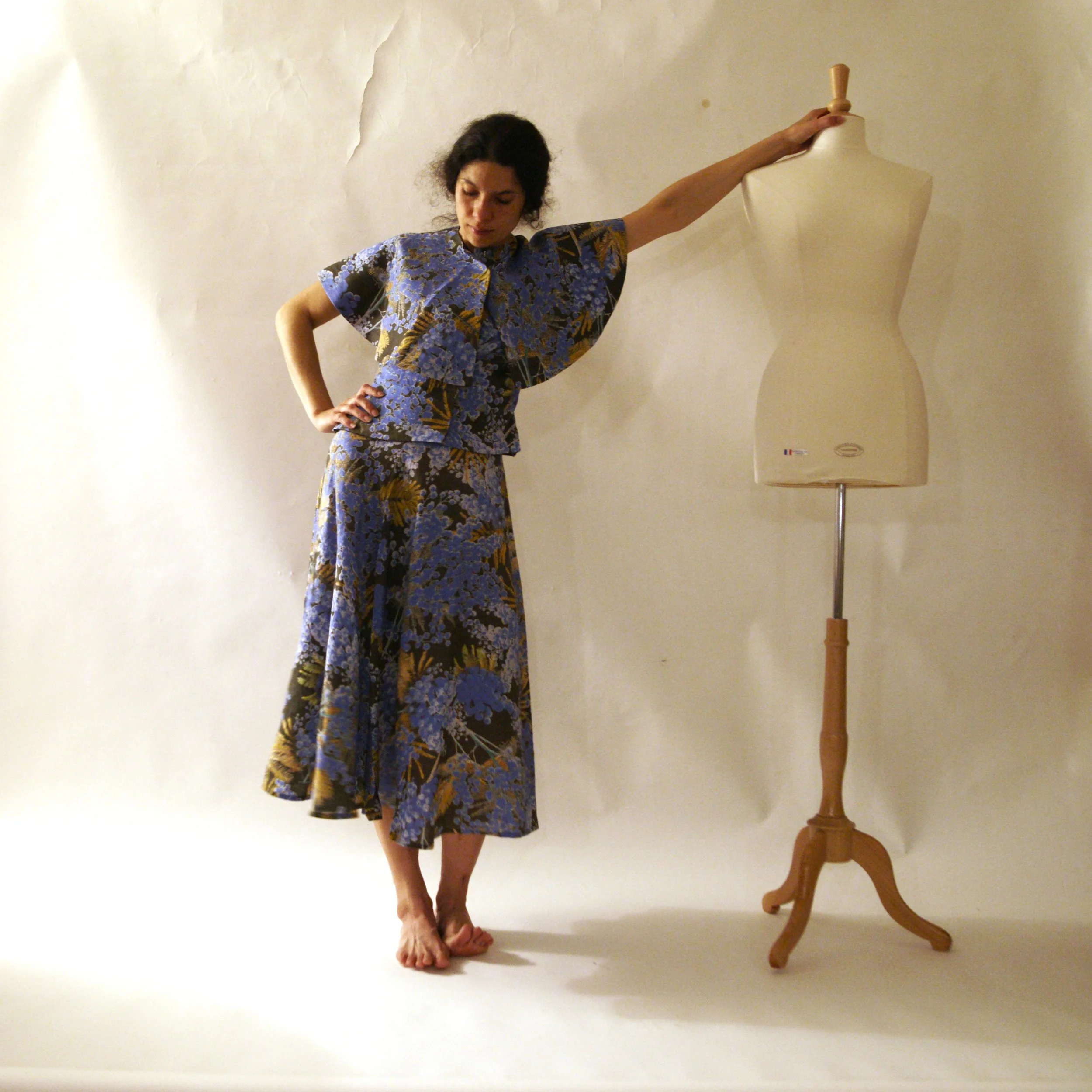 A woman in a floral dress is standing on a white floor with her eyes closed, one hand on her hip, and the other arm extended resting on a mannequin. The mannequin is on a wooden stand and is turned to the side, with a plain off-white wall in the back