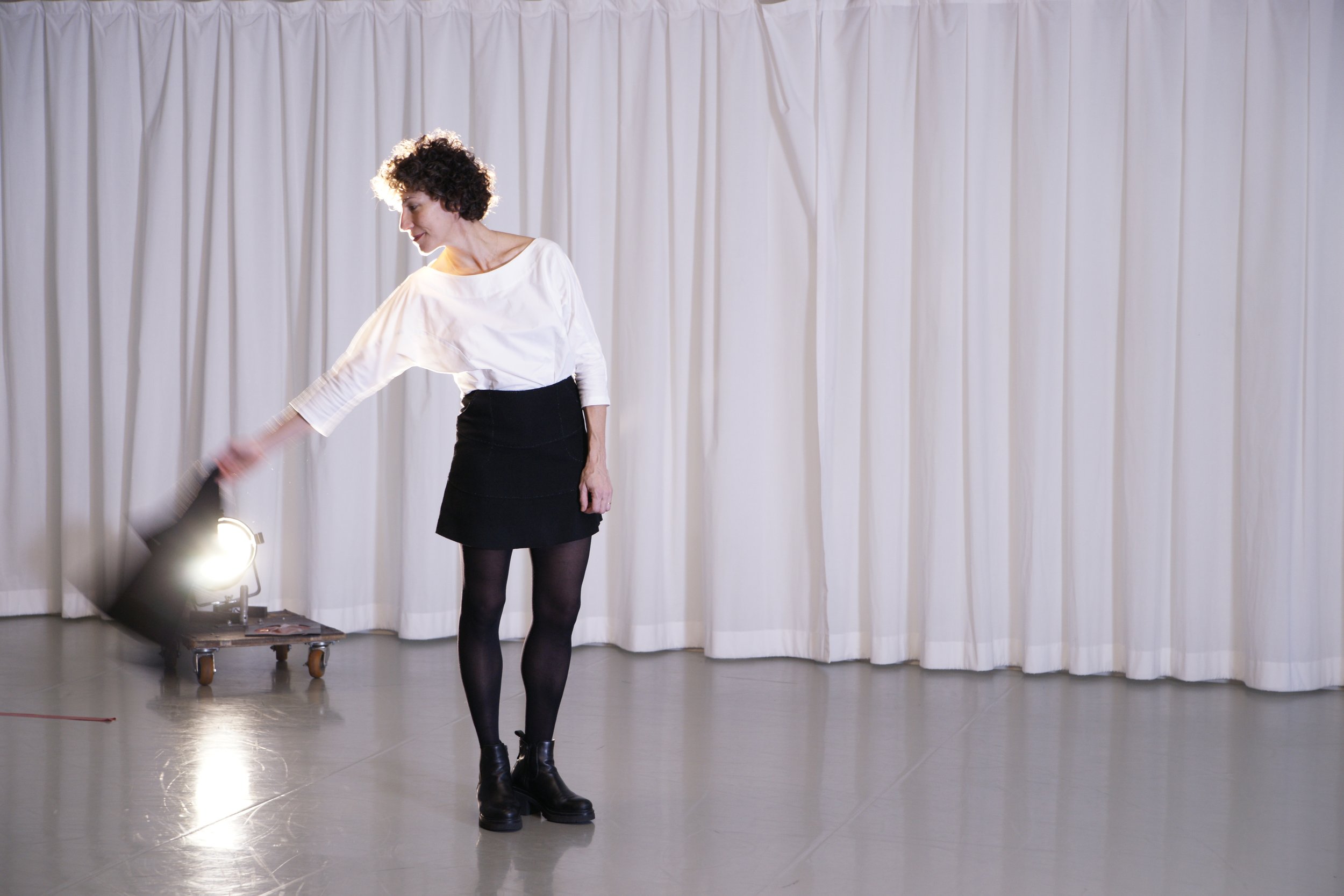 A woman with curly short hair wearing a white blouse, black skirt, and black tights, standing on a stage with a white curtain backdrop, looking down and smiling slightly.