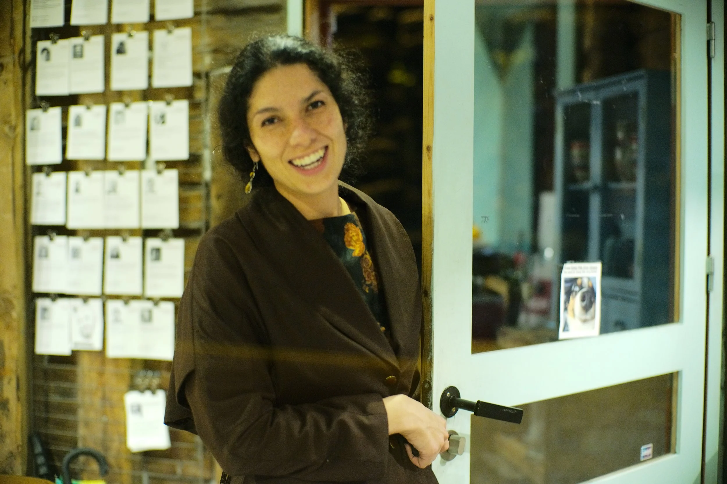 A woman with curly dark hair smiling and opening a door with a handle, standing in front of a display wall with photo cards or notes and a glass display case.