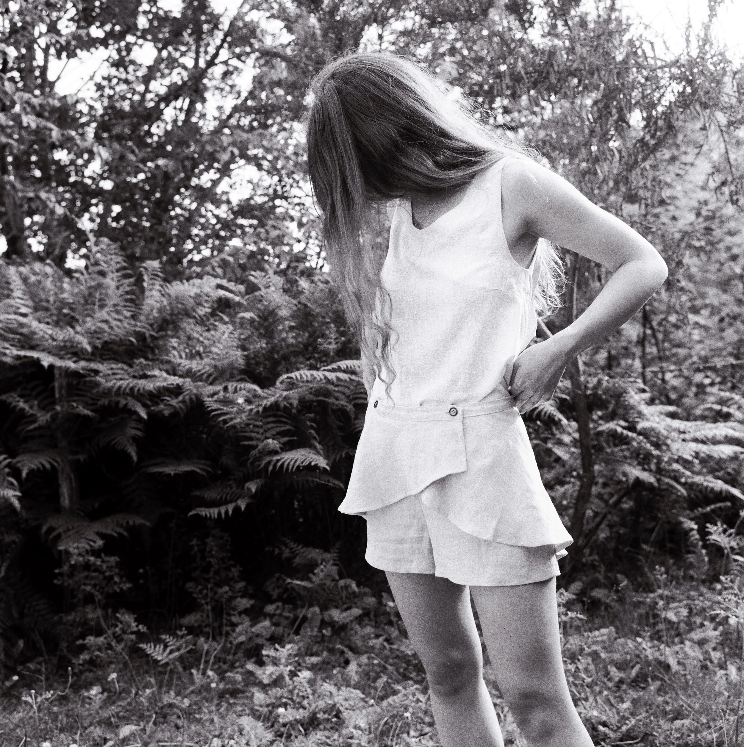 A person with long hair wearing a sleeveless top and shorts standing outdoors in a forest or wooded area with dense foliage in the background.