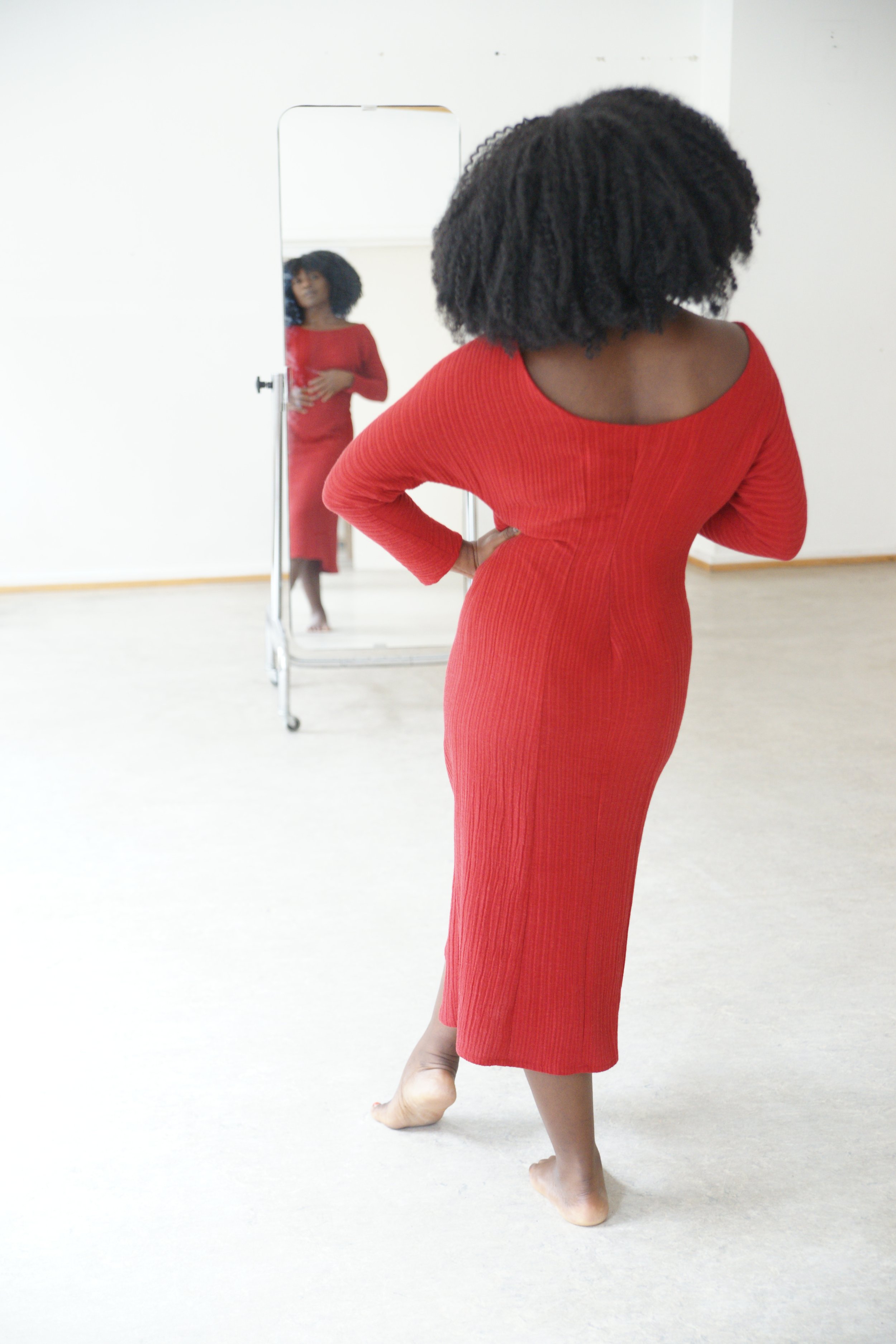 A woman in a red dress standing on one leg and looking in a mirror.