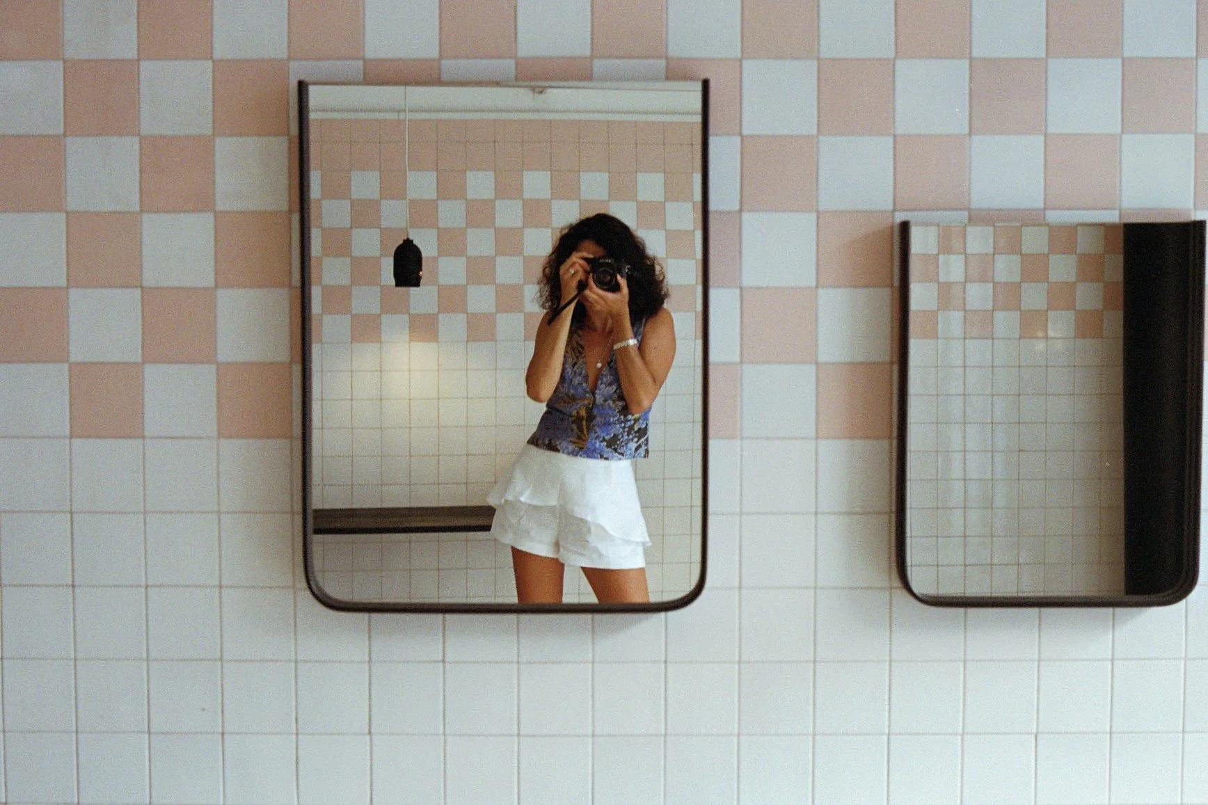 A woman taking a selfie in a mirror at a bathroom or changing room, with pink and white checkered tile walls and two mirrors, one of which she is reflected in.
