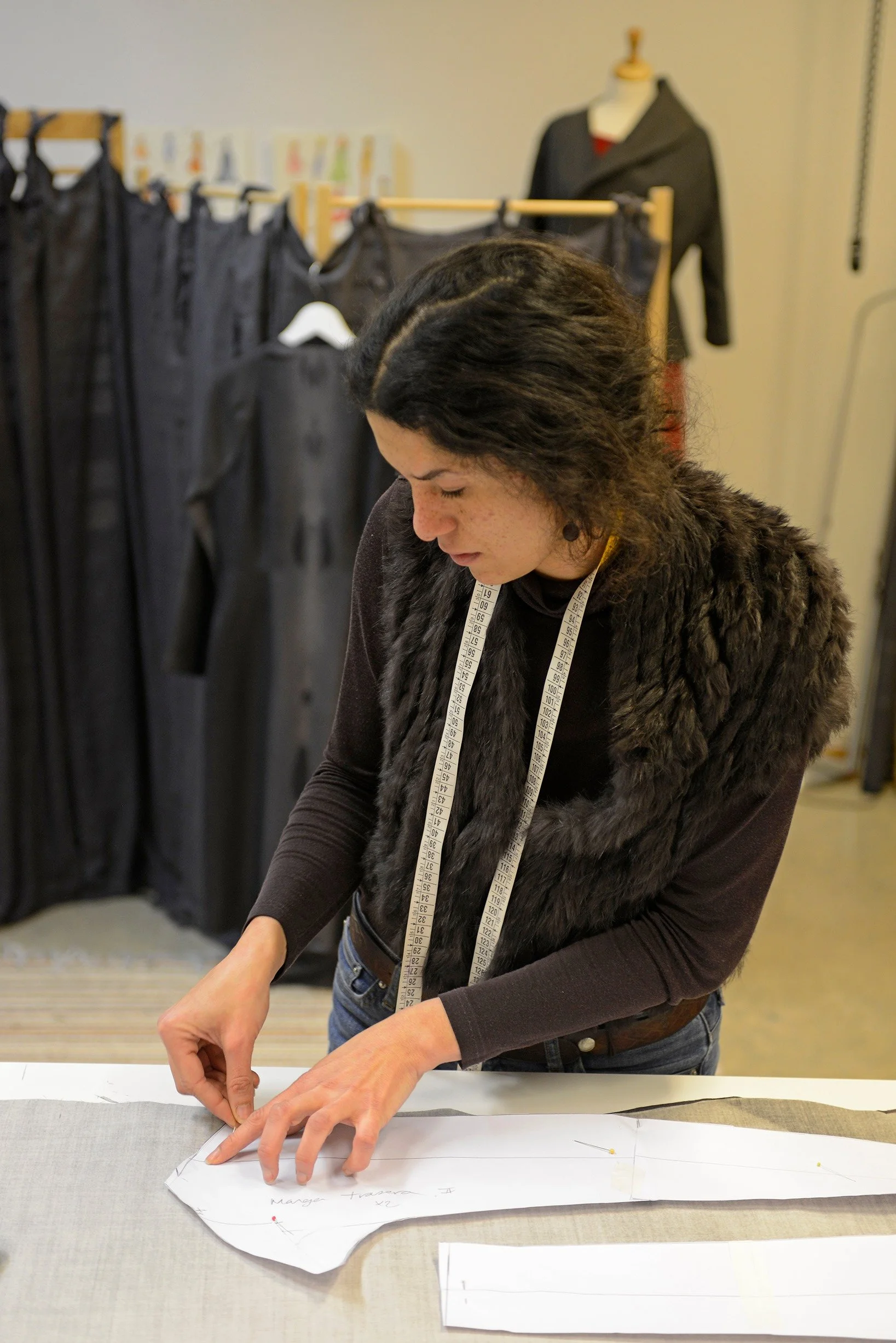 A woman with dark, wavy hair, wearing a black furry vest and a measuring tape around her neck, working on a sewing pattern at a table. In the background, there are black fabric garments hanging on racks and a mannequin dressed in dark clothing.