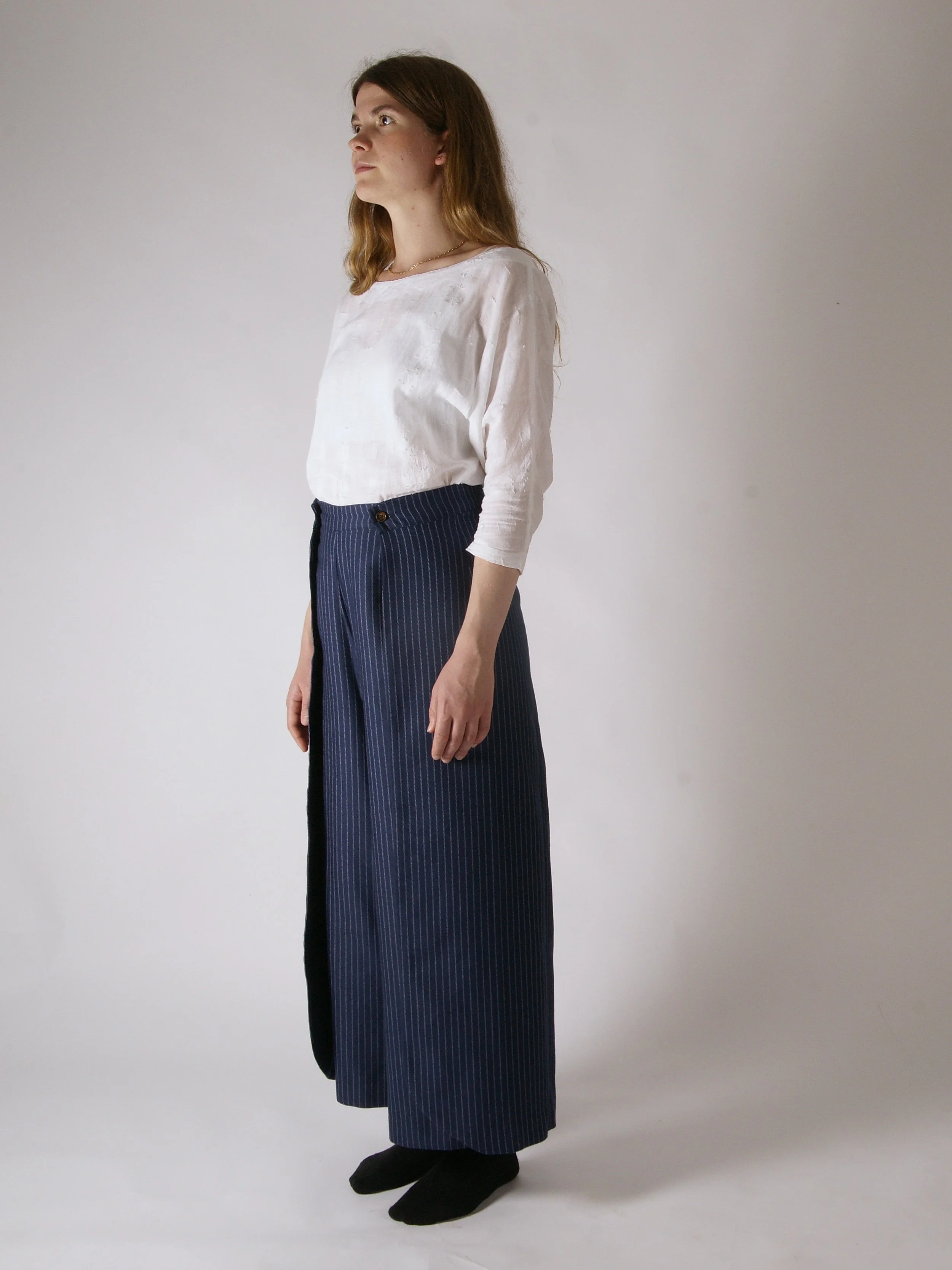 A woman with light skin and long brown hair, wearing a white long-sleeve shirt, a dark blue pinstripe maxi skirt, and black ankle boots, standing against a plain gray background.