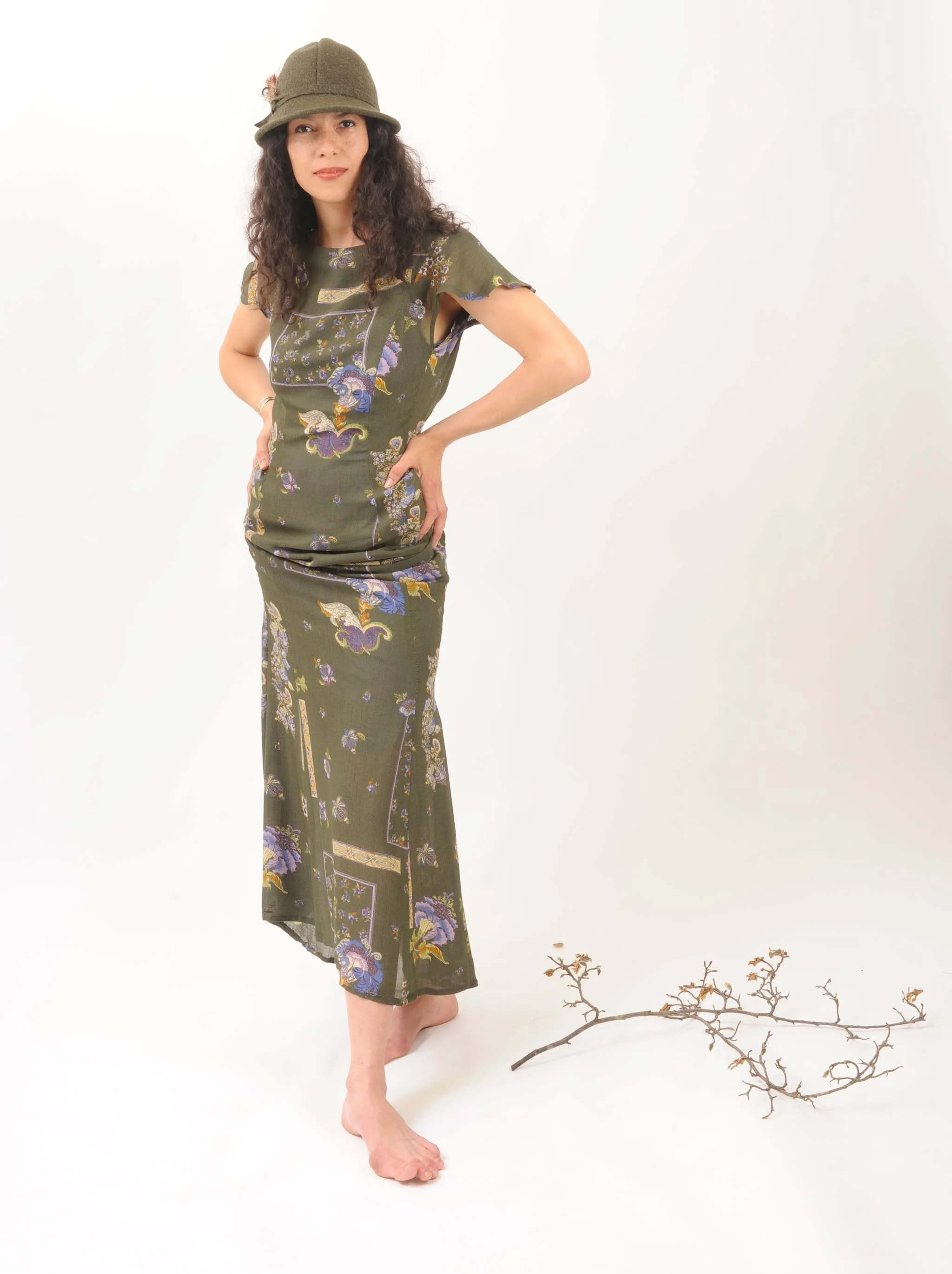 A woman with curly dark hair wearing a green hat and an olive-green floral dress, standing barefoot next to a small branch with dried twigs on a white background.