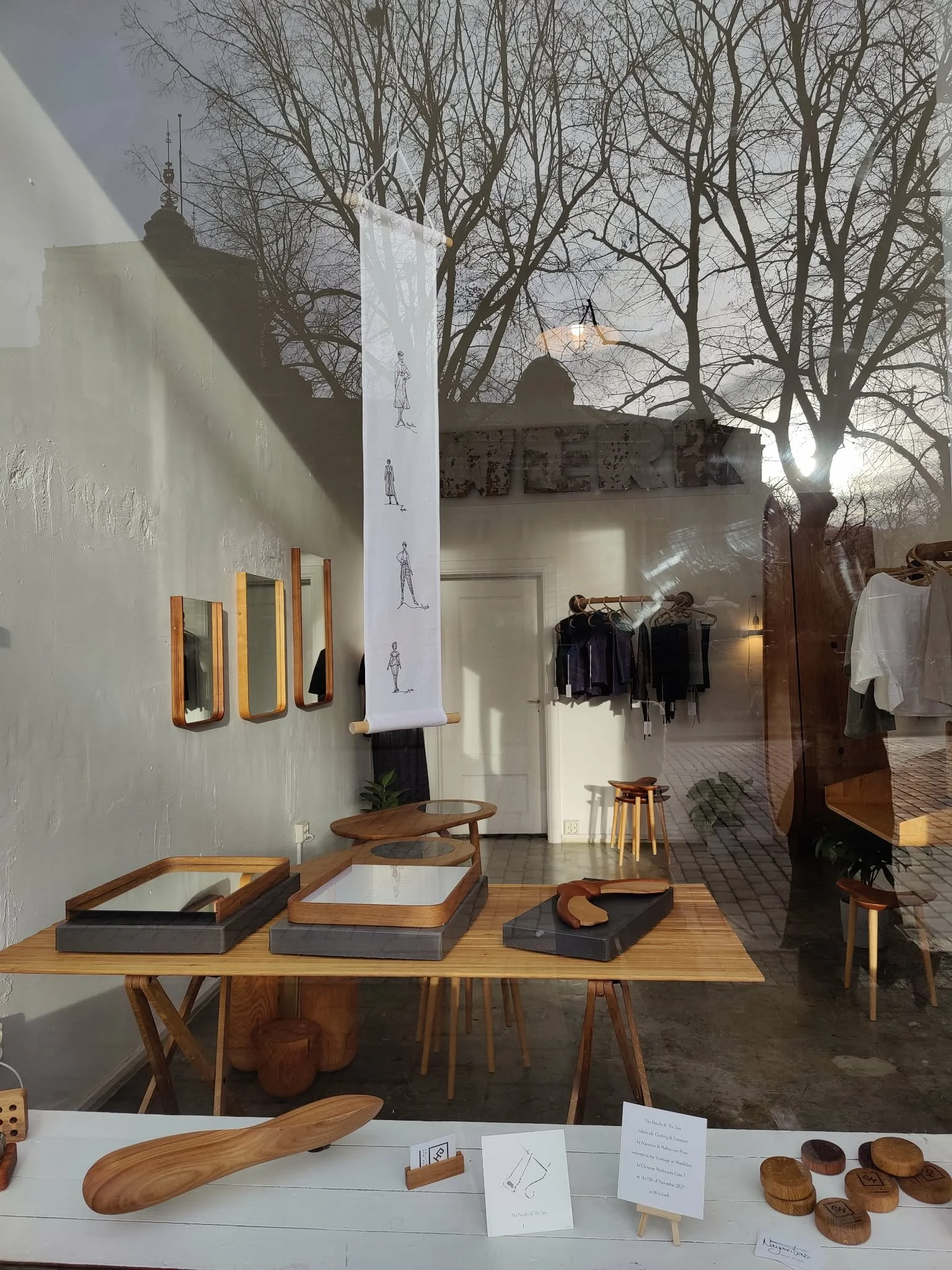 View through a shop window featuring wooden furniture and decorative items, with a reflection of leafless trees and a historic building outside.