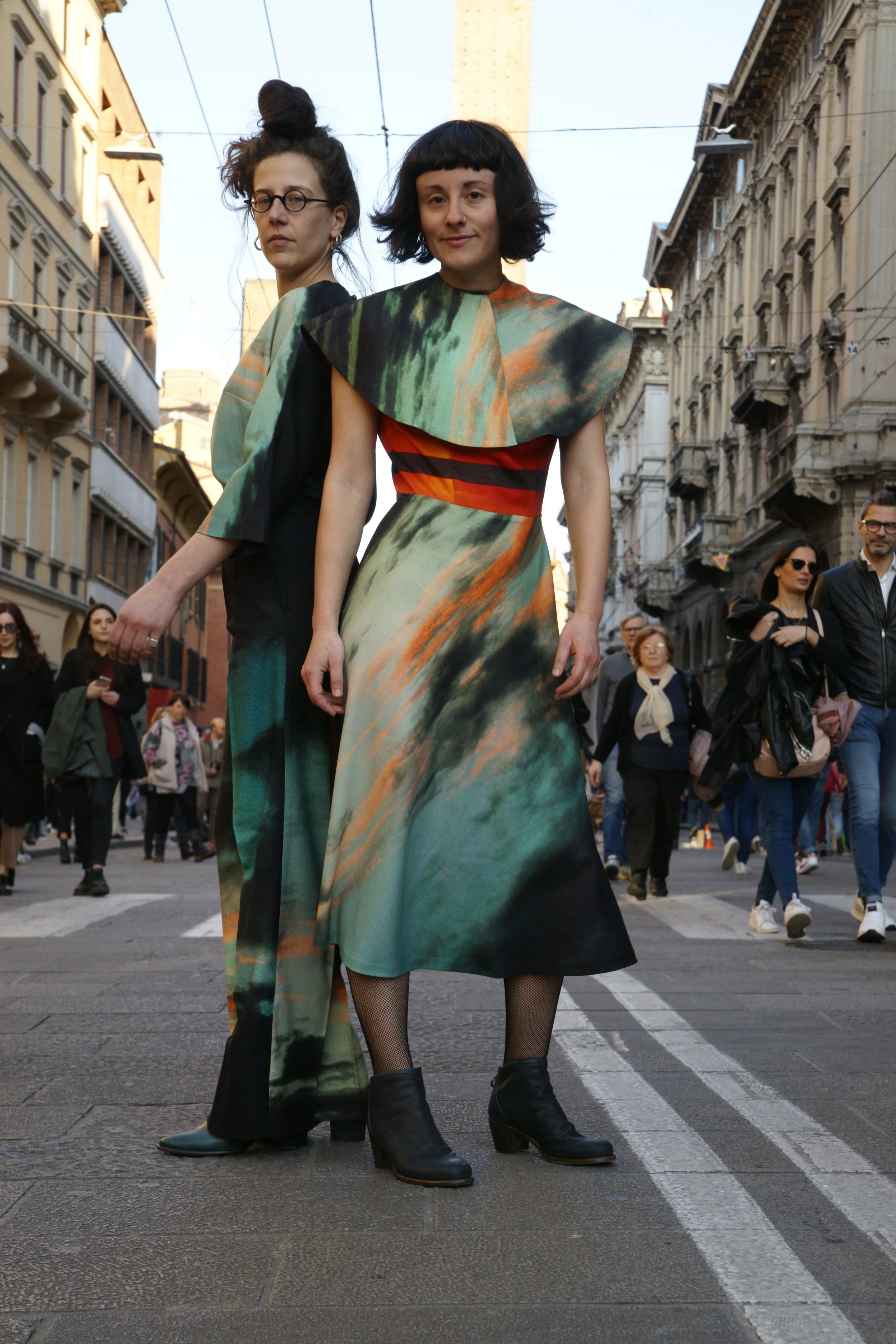 Two women standing in the street during an outdoor event or parade, dressed in flowy, colorful, abstract-patterned dresses with black boots, surrounded by onlookers and buildings.