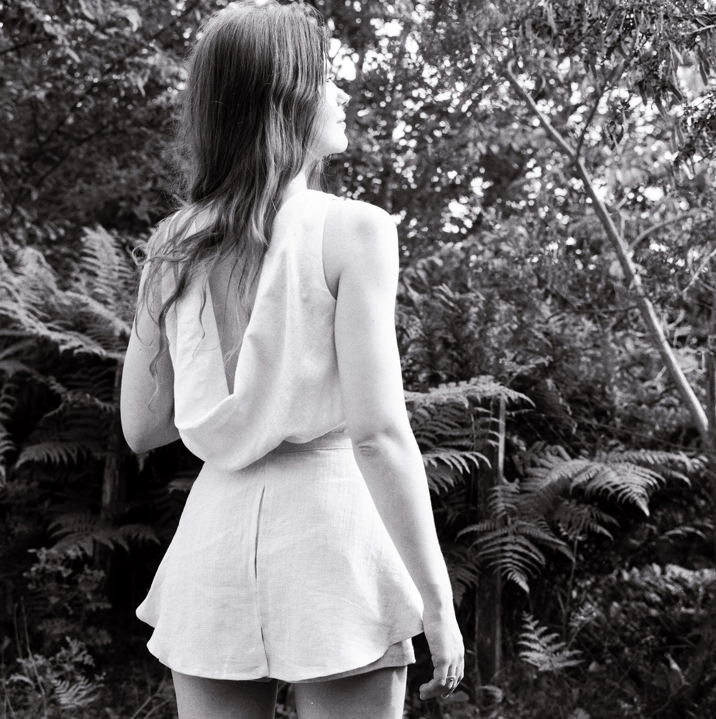 A woman with long hair facing sideways outdoors among trees and foliage, wearing a sleeveless top and shorts.