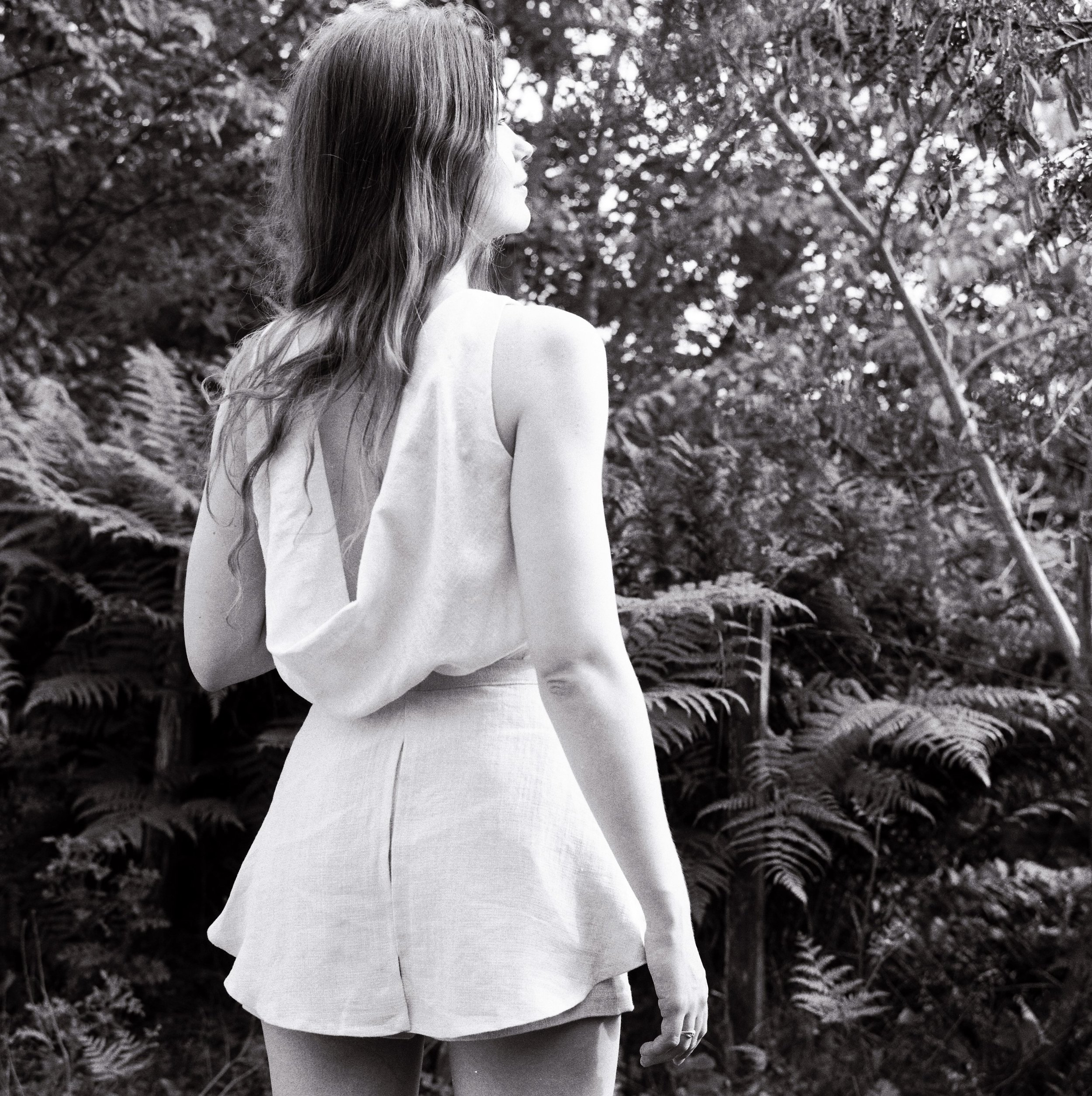 A woman with long hair standing outdoors among trees, facing sideways, wearing a sleeveless top.