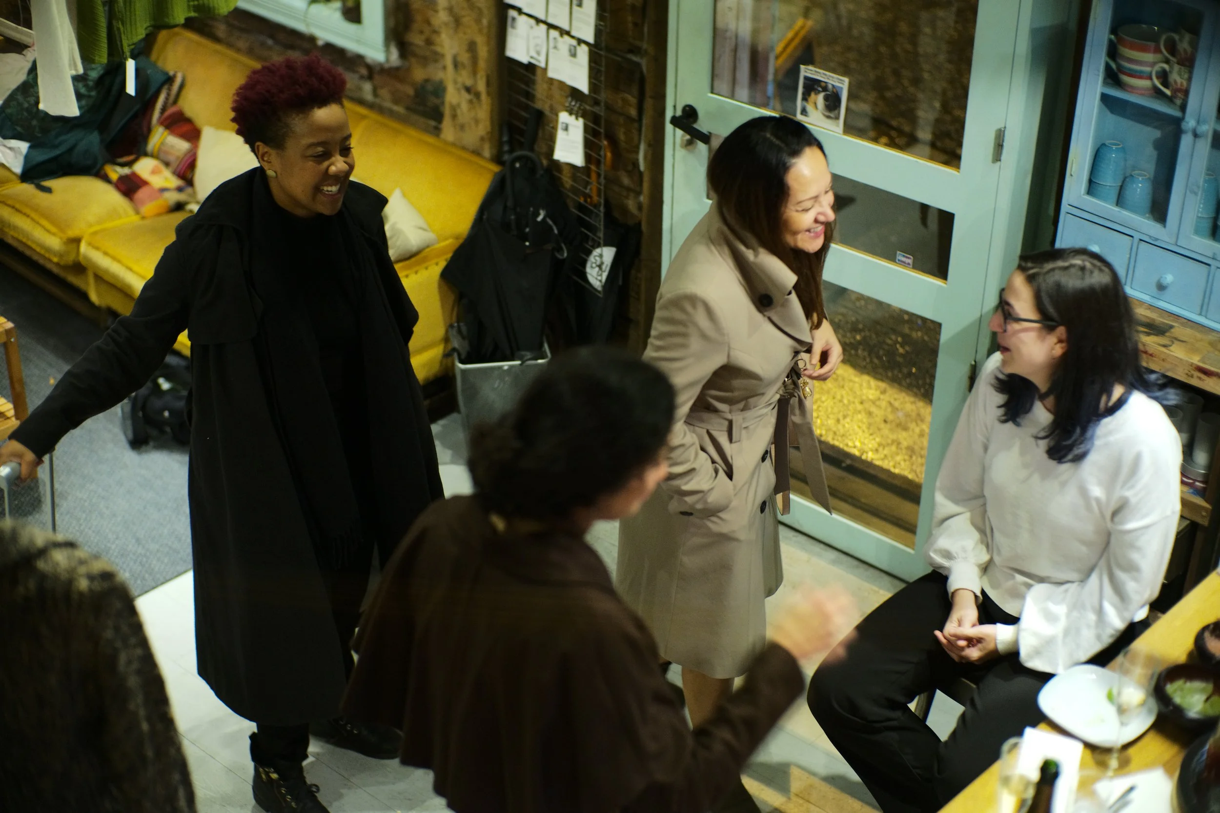 A group of people, mostly women, are gathered indoors, smiling and engaging in conversation around a table with food, in a cozy, rustic space with wooden walls and a blue cabinet.