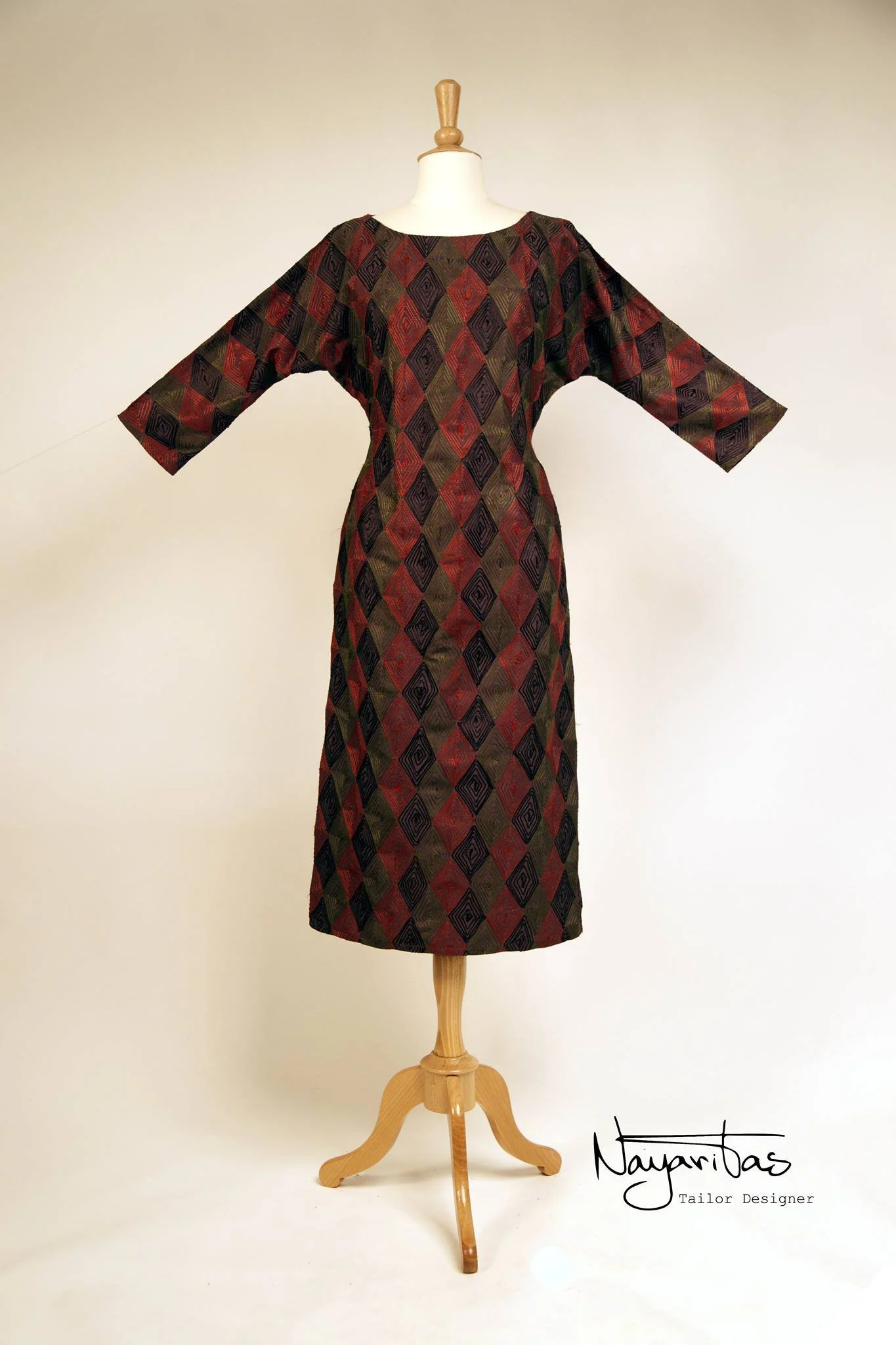 A long dress with a geometric diamond pattern in shades of red, black, and green displayed on a mannequin. The dress has three-quarter sleeves and a round neckline, and is made of a shiny fabric. The mannequin is on a wooden stand, and the background