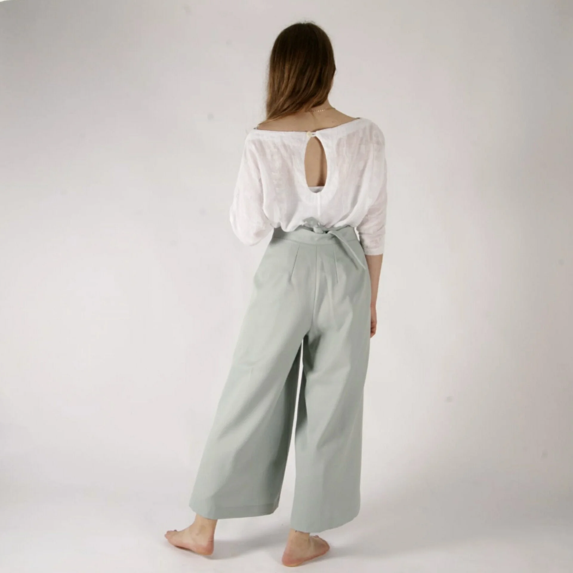 A woman with brown hair standing barefoot on a white background, wearing a white top with a keyhole back and gray wide-leg pants.