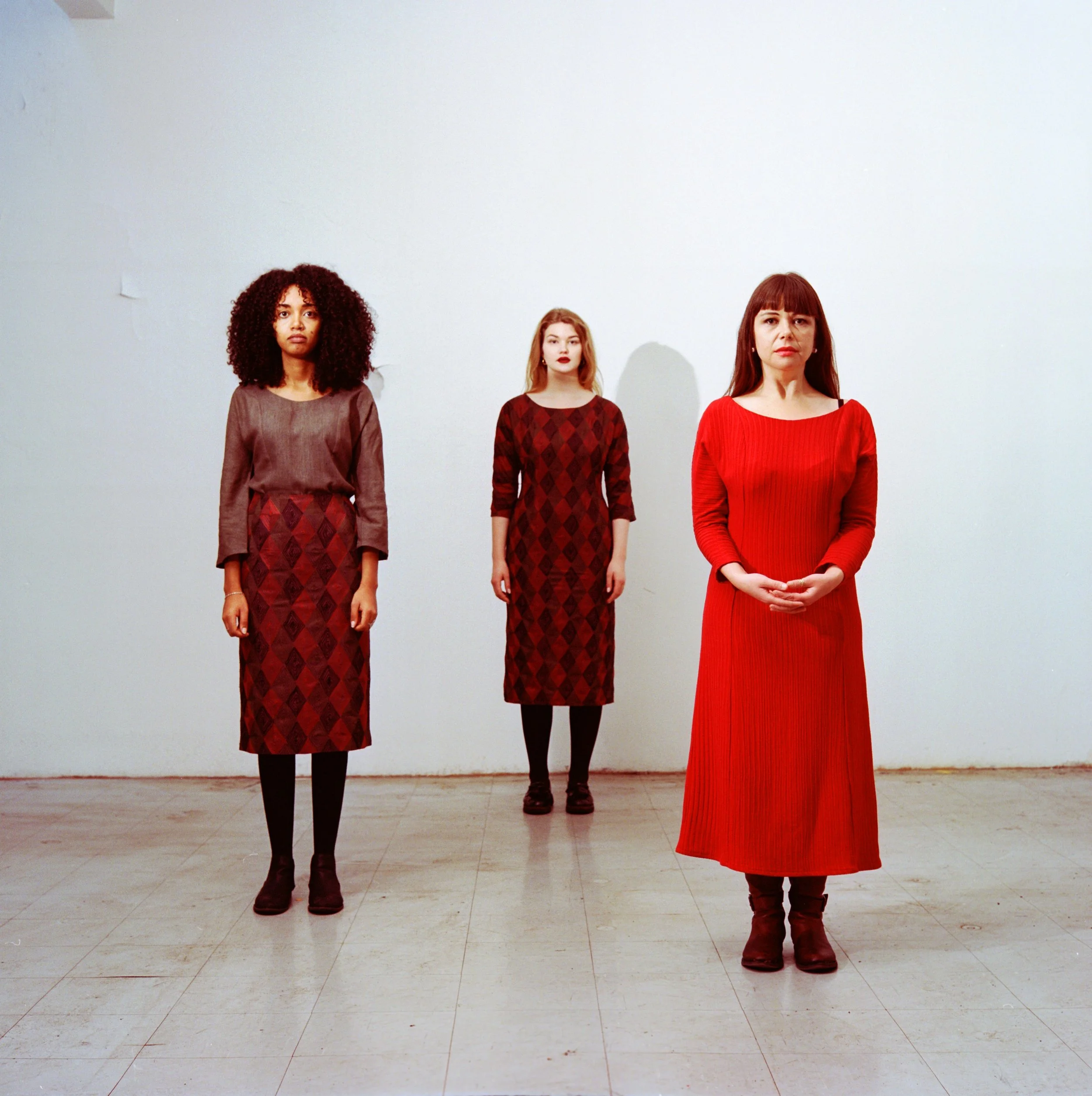 Three women standing in a line with a plain light-colored wall behind them, dressed in shades of red and patterned clothing.