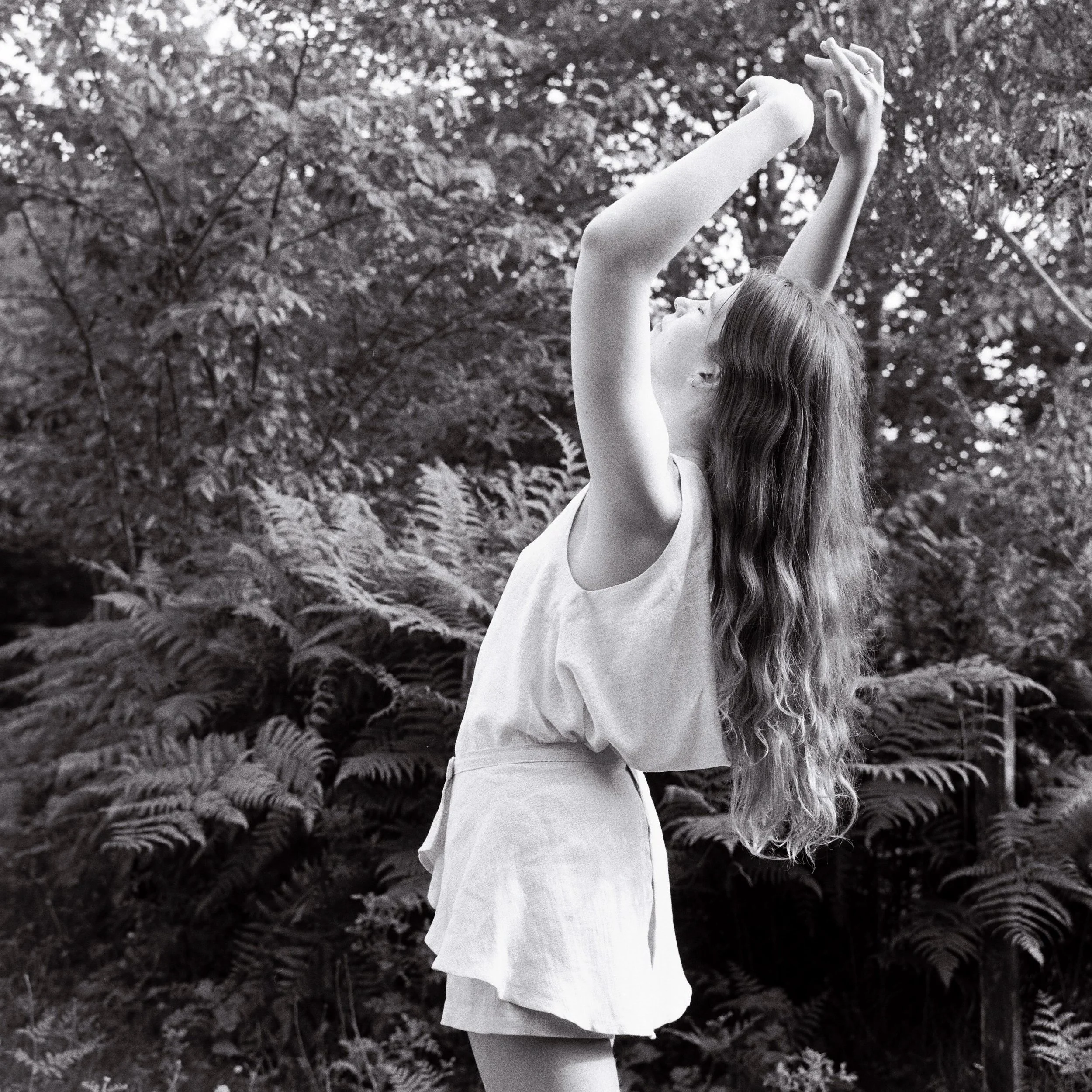 A woman with long hair wearing a sleeveless top and skirt, outdoors among foliage, stretching her arms upward and clasping her hands together.