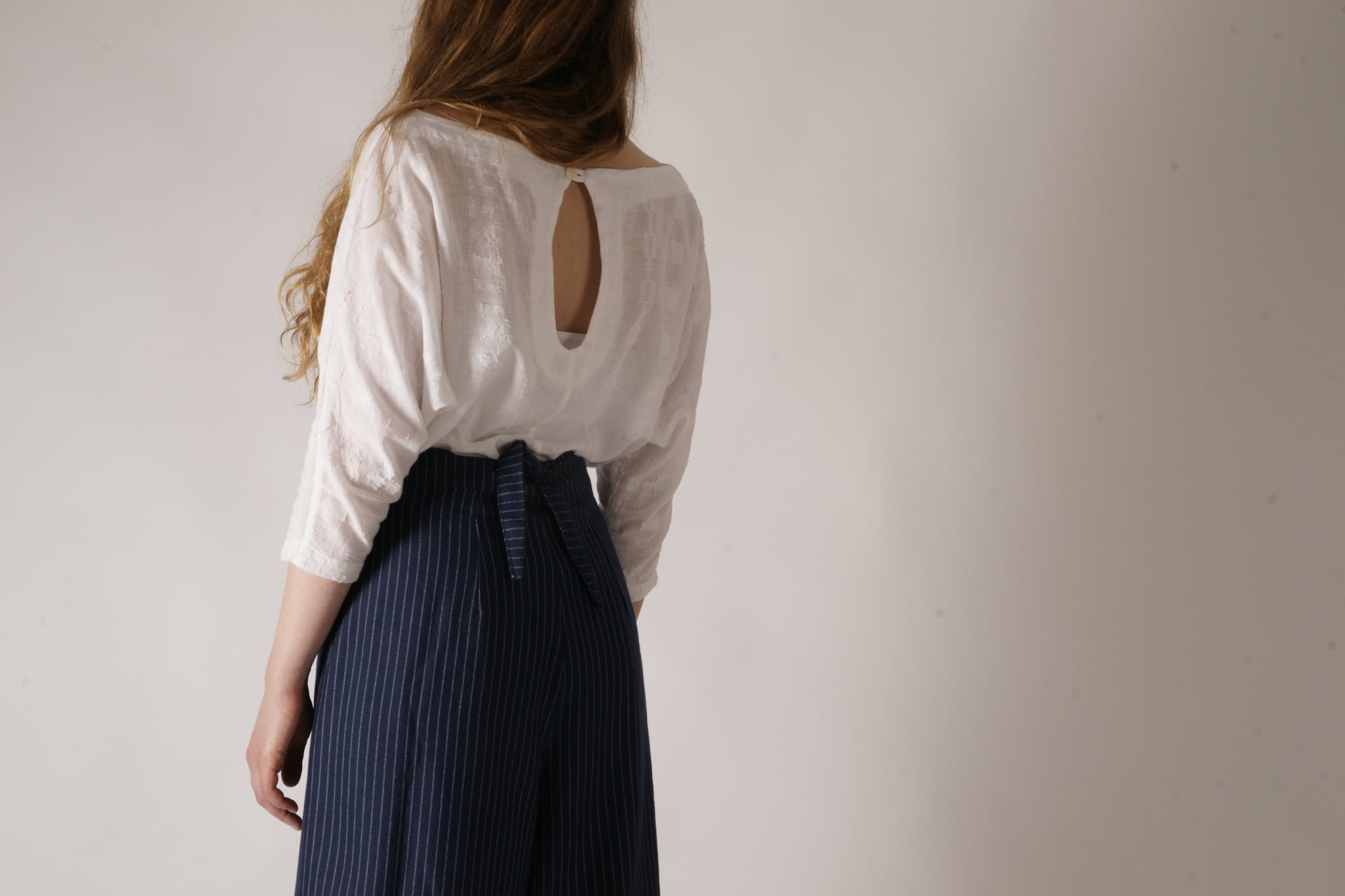A woman with long reddish hair wearing a white blouse with a keyhole opening on the back and navy pinstripe pants against a plain white wall.