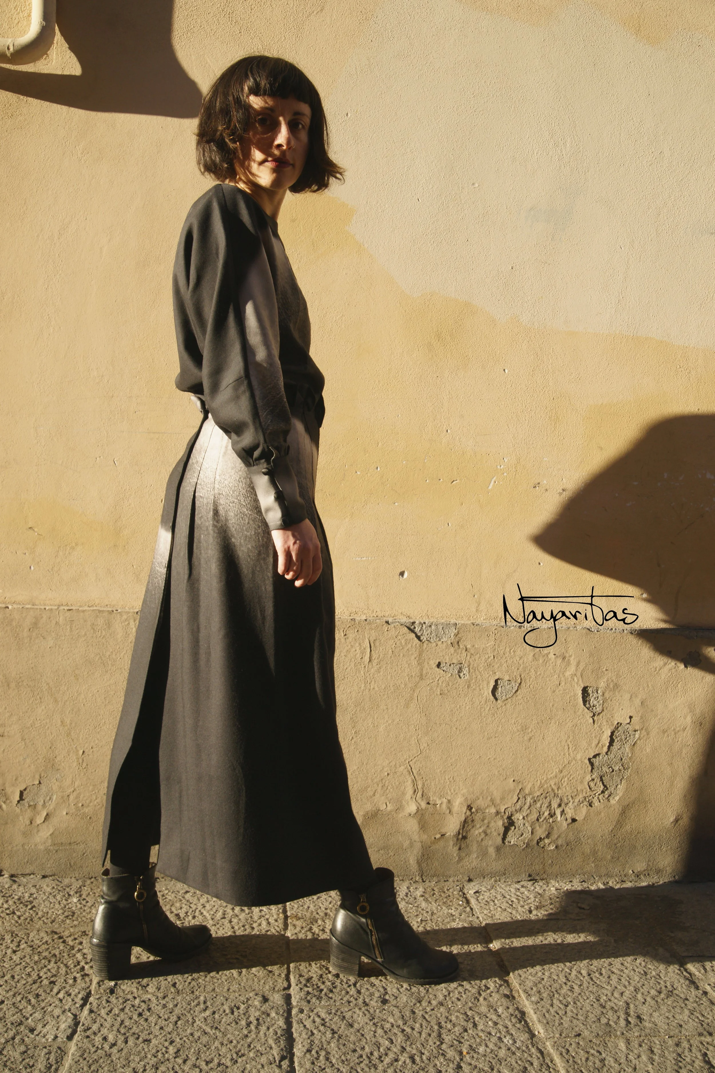 A woman with short dark hair wearing a dark long-sleeve top, wide-leg pants, and black boots with high heels, standing against a beige textured wall; sunlight casting shadows.