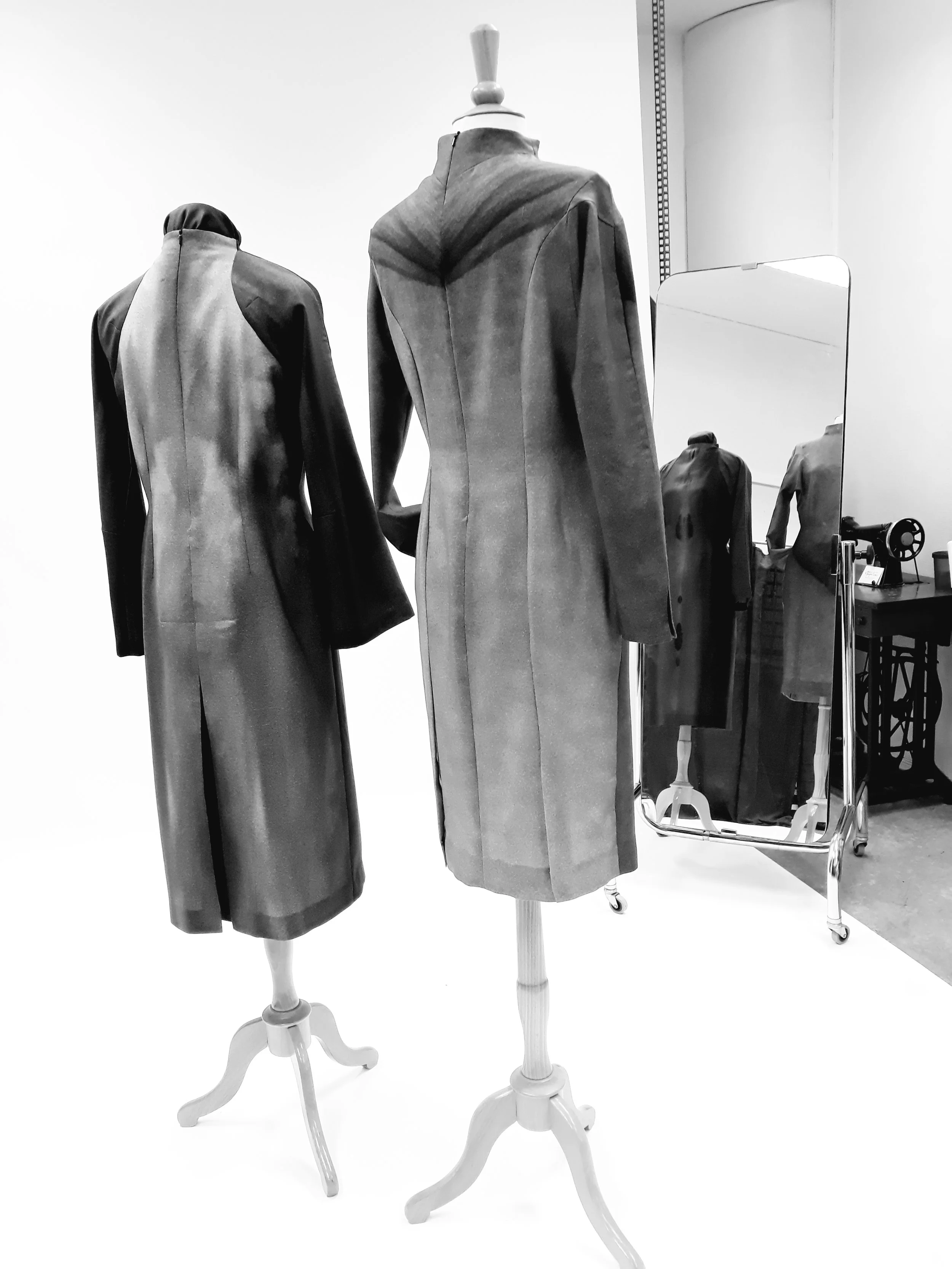 Black and white photo of two vintage mannequins on white wooden stands, one with a long coat and the other with a suit in a dressing room or store setting, with a mirror and a sewing machine in the background.