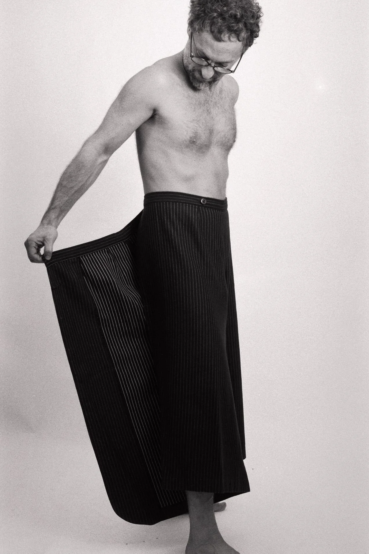 A shirtless man with curly hair and glasses, wearing pinstriped wide-leg pants, is standing against a plain background, holding the hem of his pants with his left hand.